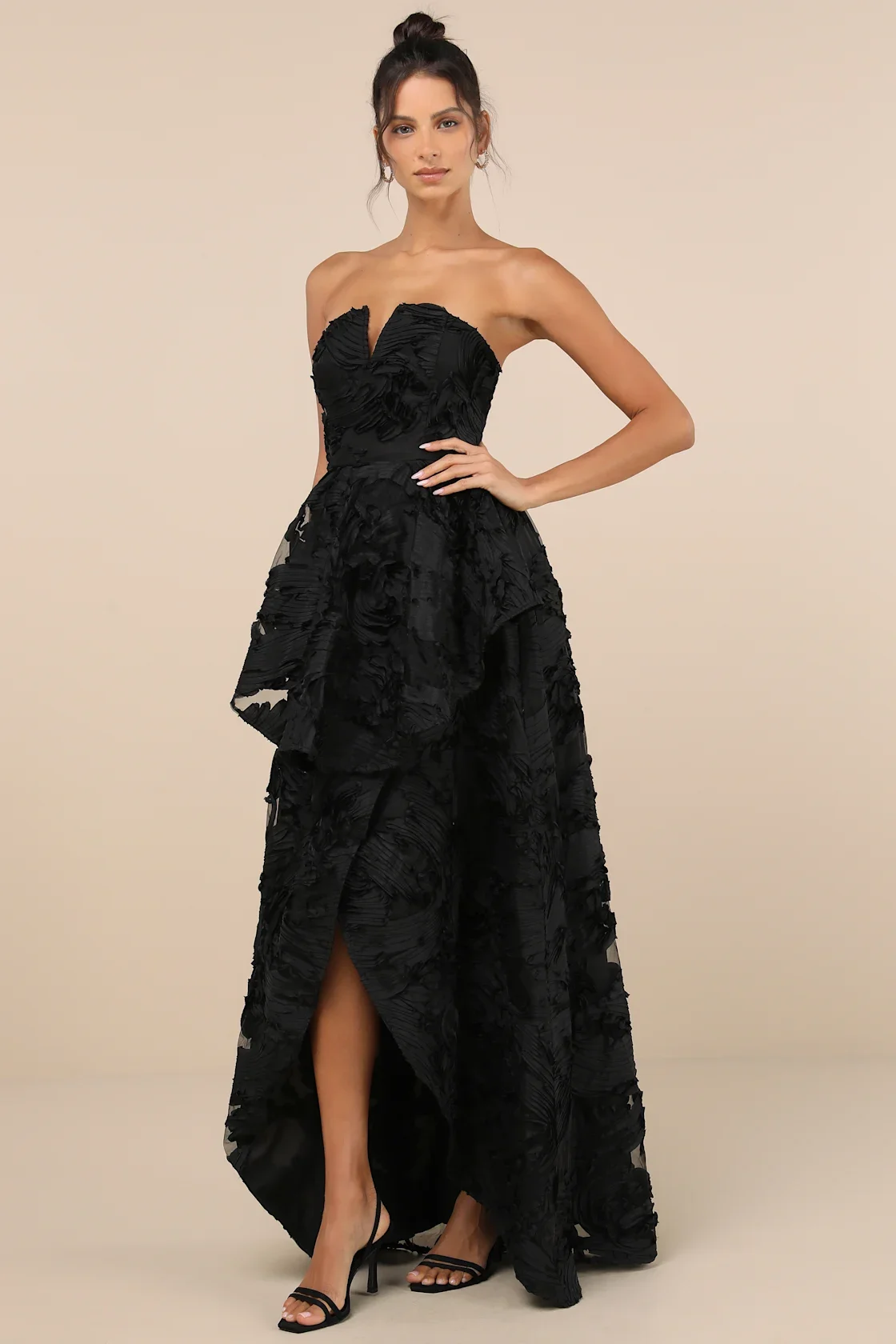 A woman wearing a strapless black gown with textured floral-like details and a high slit, standing against a plain beige background.