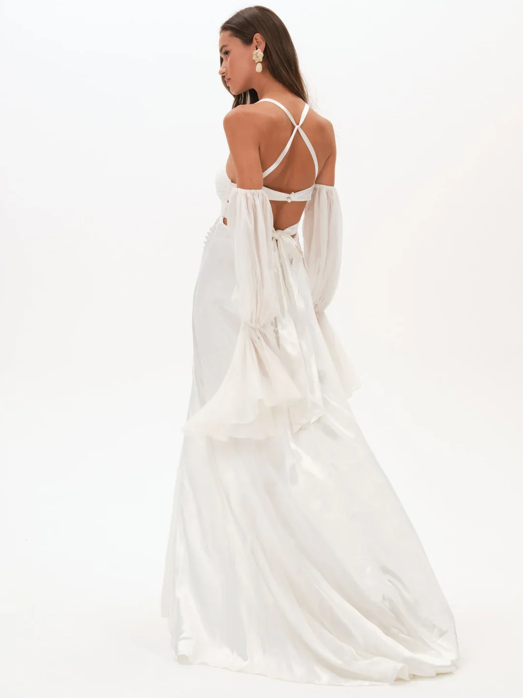 A woman in a white, backless dress with puffed, long sleeves and ruffled details, standing against a plain white background.