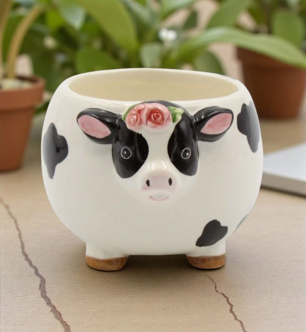 Ceramic planter shaped like a cow with a painted face and pink flower on its head, sitting on a surface with plants and pots in the background.