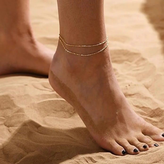 Close-up of a woman's foot and ankle on sandy ground, with delicate anklets and painted toenails.