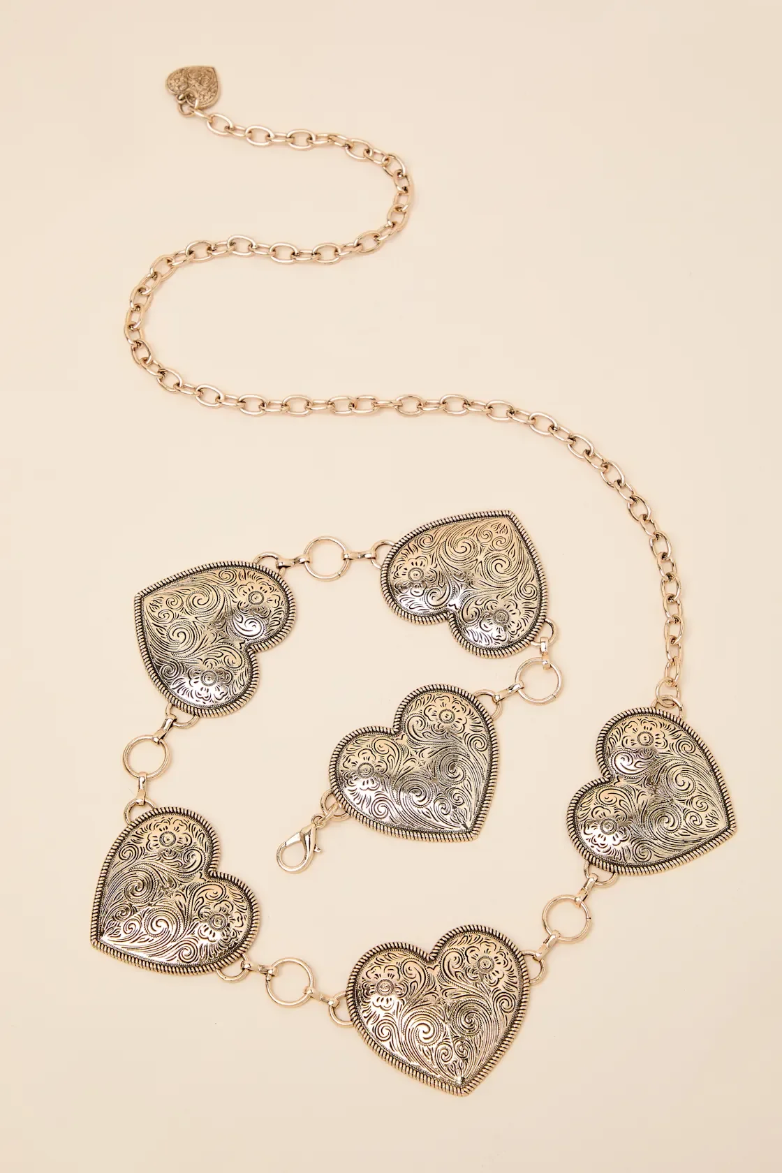 Silver heart-shaped jewelry with intricate swirl and floral engravings, interconnected with small rings on a light beige background.