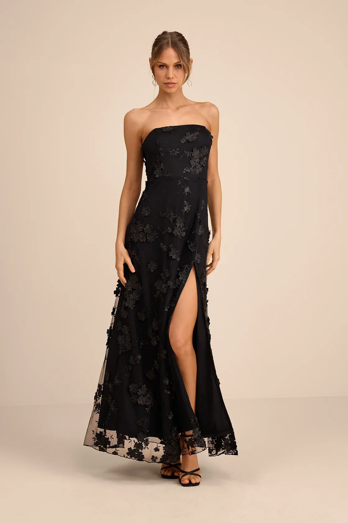 A woman wearing a black strapless dress with floral embroidery and a high slit, standing against a plain beige background.
