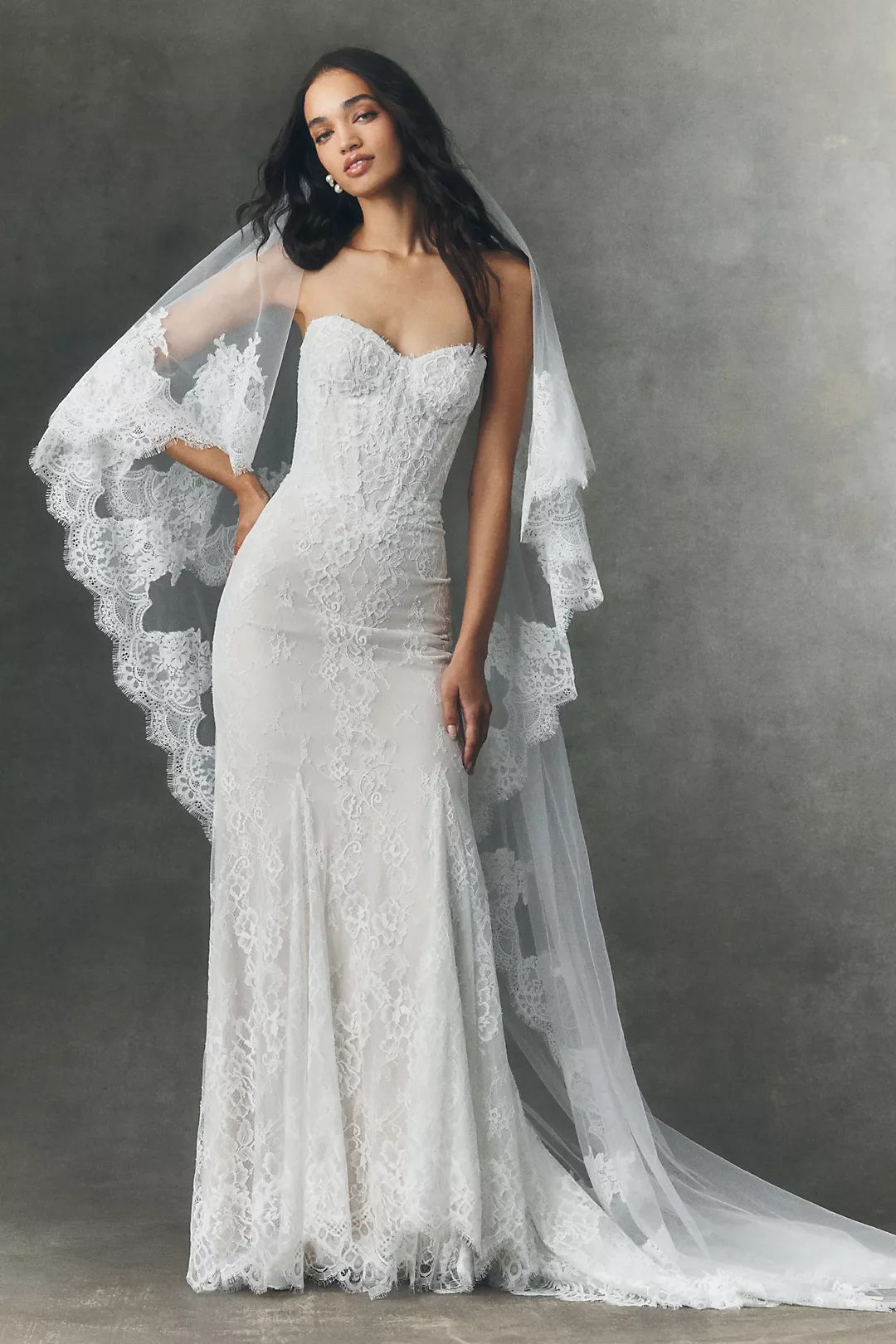 A woman wearing a strapless white lace wedding gown with a long train and lace cape, standing against a plain gray background.