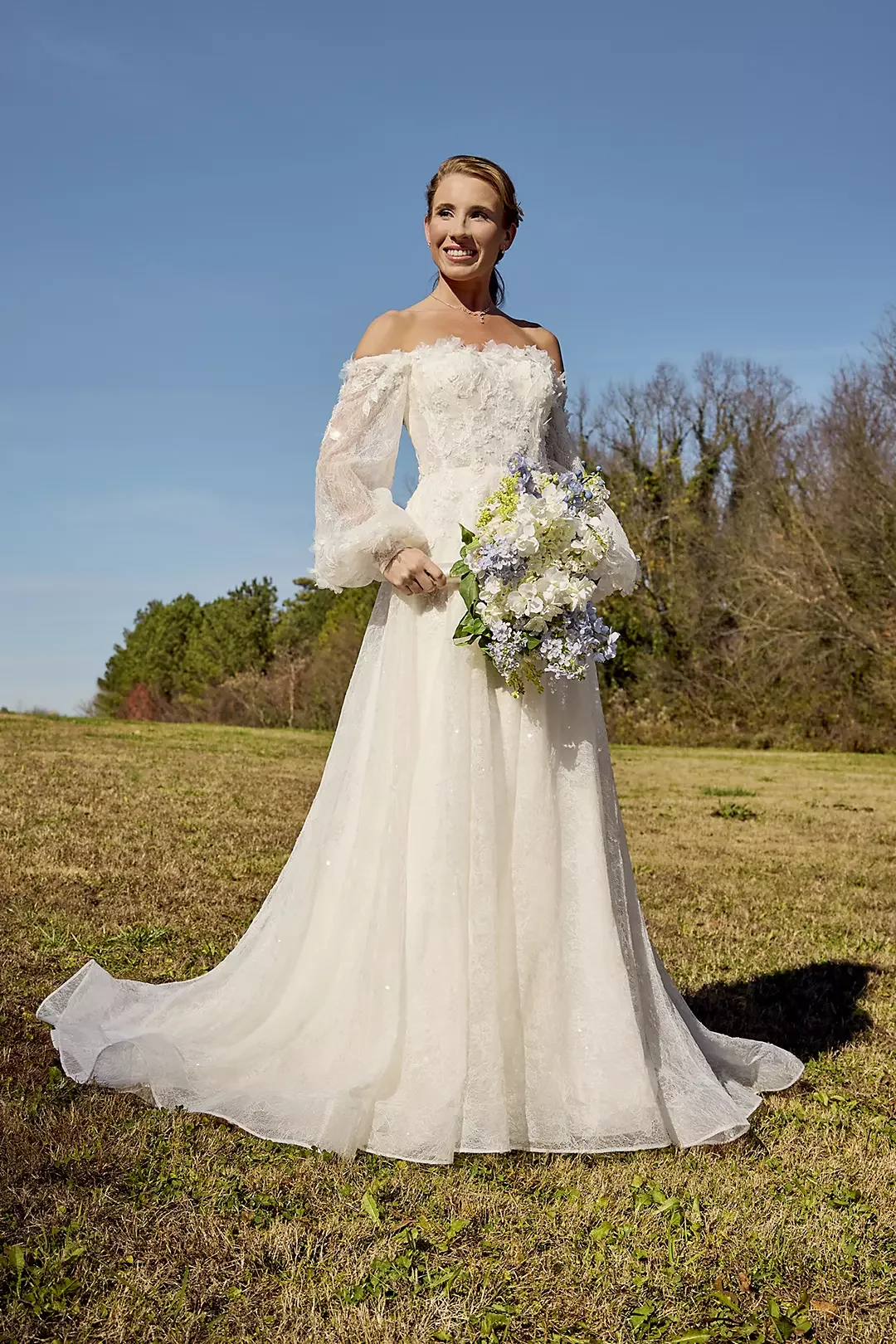A woman in a white wedding dress holding a bouquet of white and purple flowers outdoors on a sunny day with trees and a blue sky in the background.