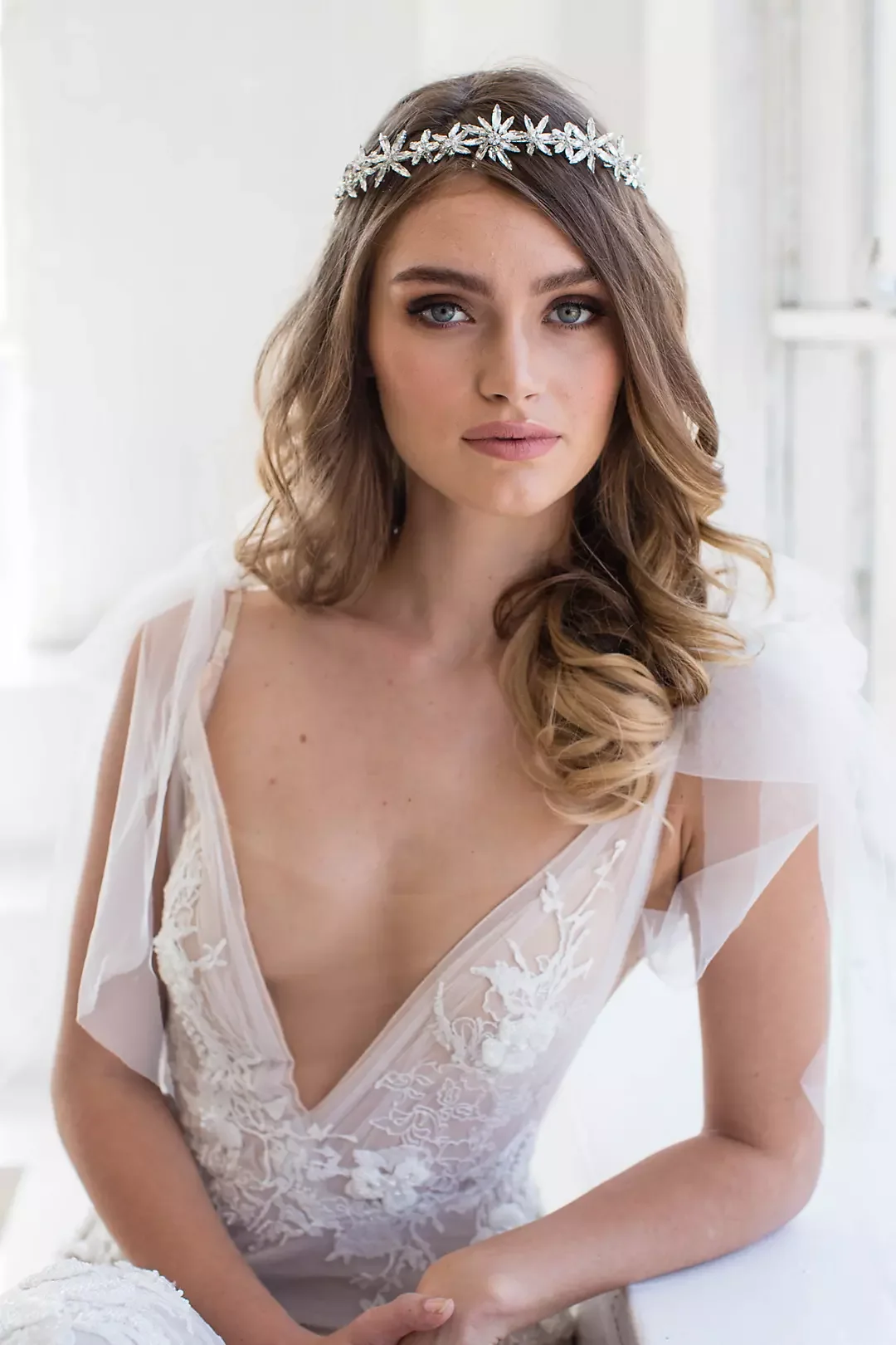 A young woman with long wavy brown hair, wearing a floral headband, a white lace dress, and natural makeup, sitting indoors near a window.
