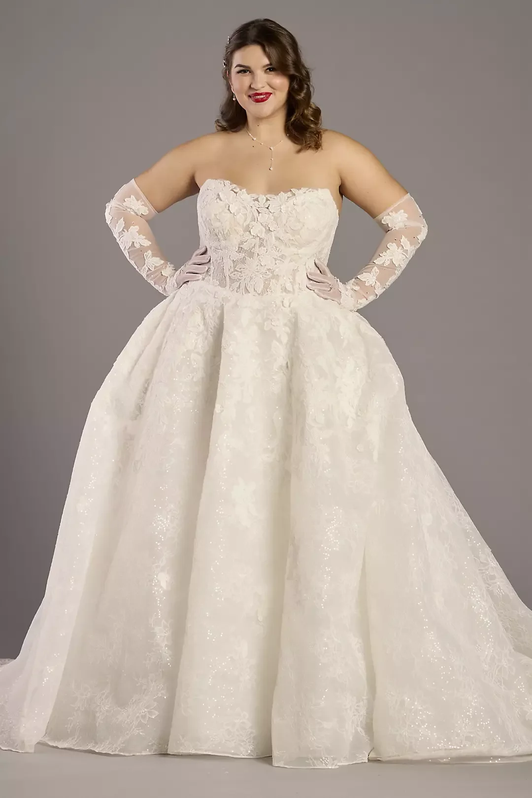 A woman in a strapless white wedding gown with lace details and long sheer lace gloves, standing against a neutral background.