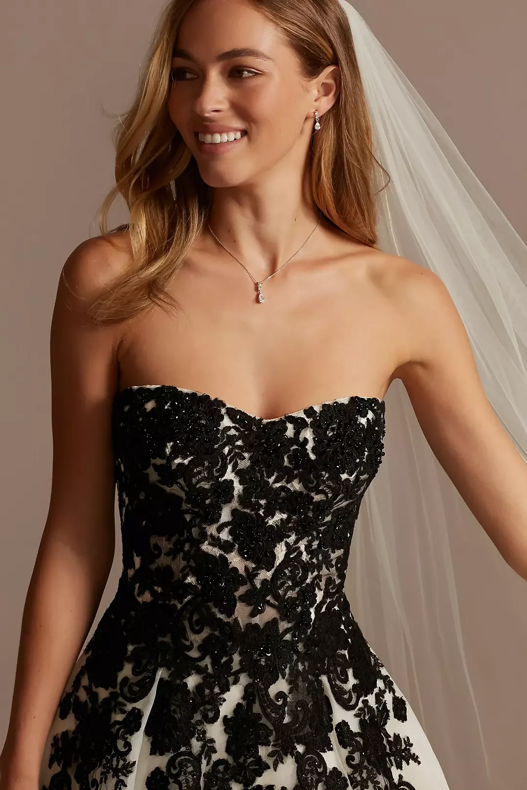 A woman in a wedding dress with black lace embroidery, a strapless neckline, a veil, and jewelry including earrings and a necklace, smiling and looking to the side.