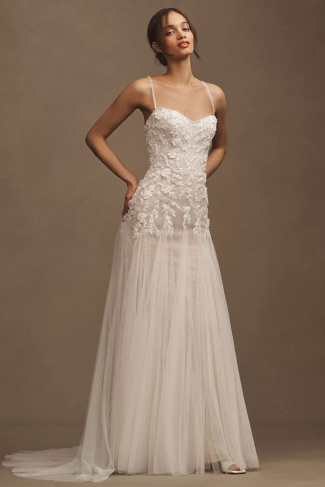 A woman wearing a white wedding dress with lace floral embroidery on the bodice, thin straps, and a flowing tulle skirt.