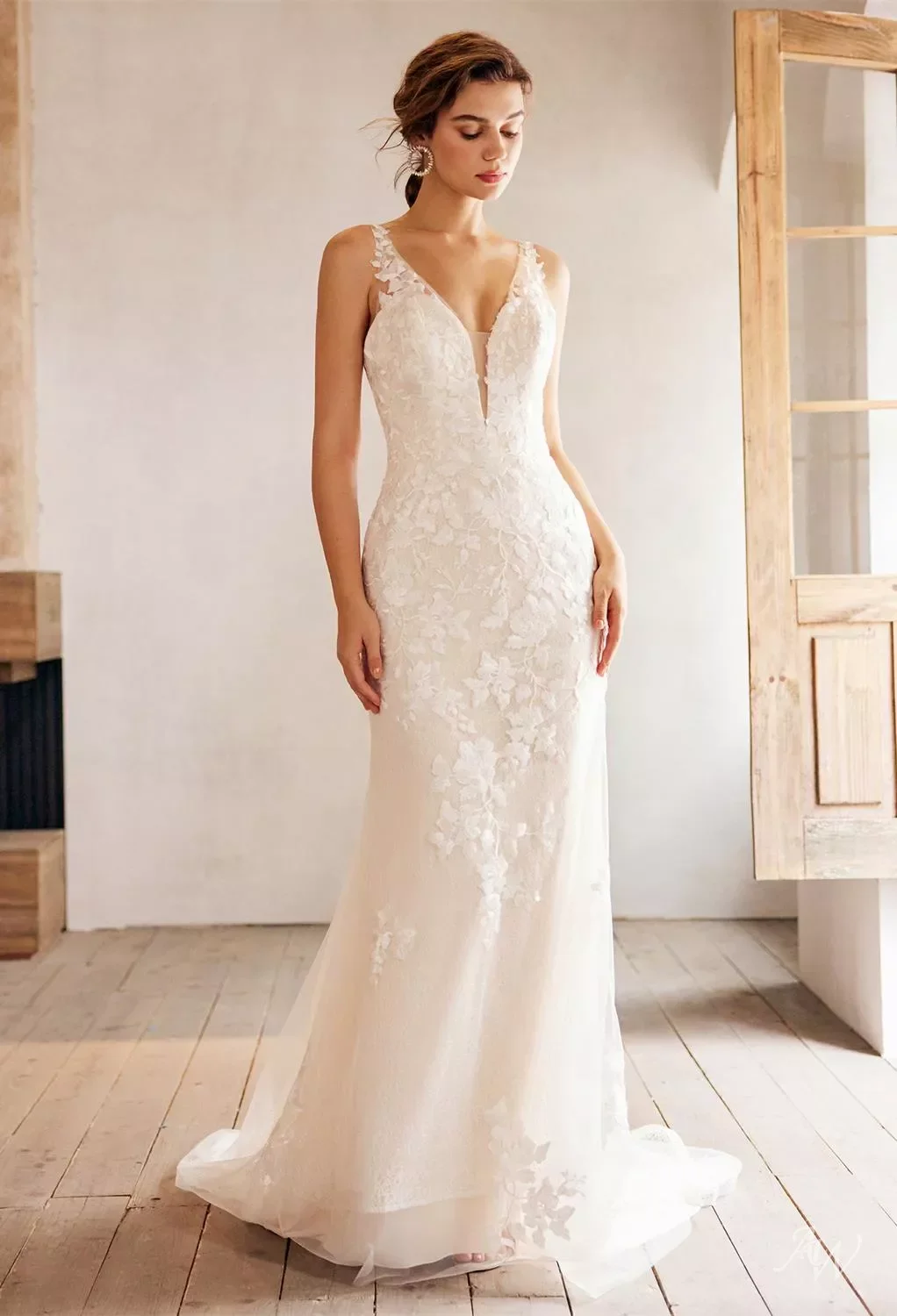 A woman wearing a white lace wedding gown with floral details and a plunging neckline, standing indoors near a wooden window frame.
