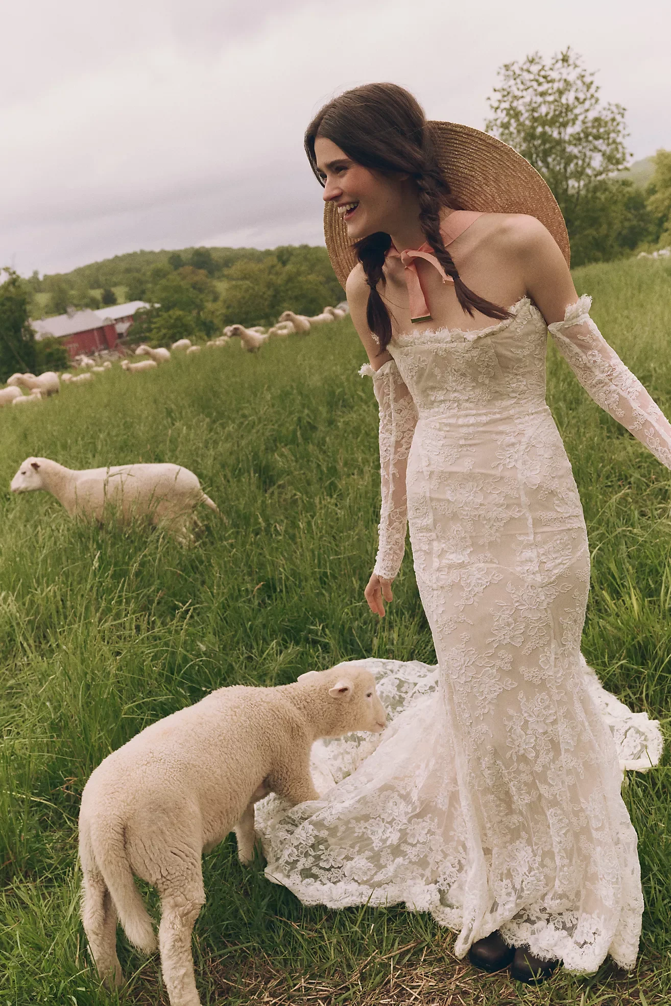 A woman in a wedding dress with long lace sleeves and a large straw hat standing in a grassy field with sheep, some grazing and some lying down, around her.