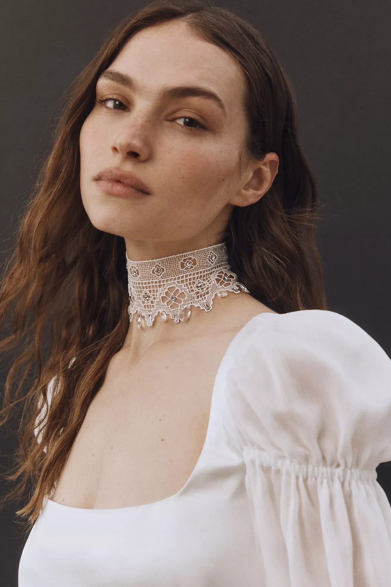 A young woman with long, wavy brown hair and fair skin wearing a white top with puffed sleeves and an intricate lace choker. She has natural makeup and is standing against a plain dark background.