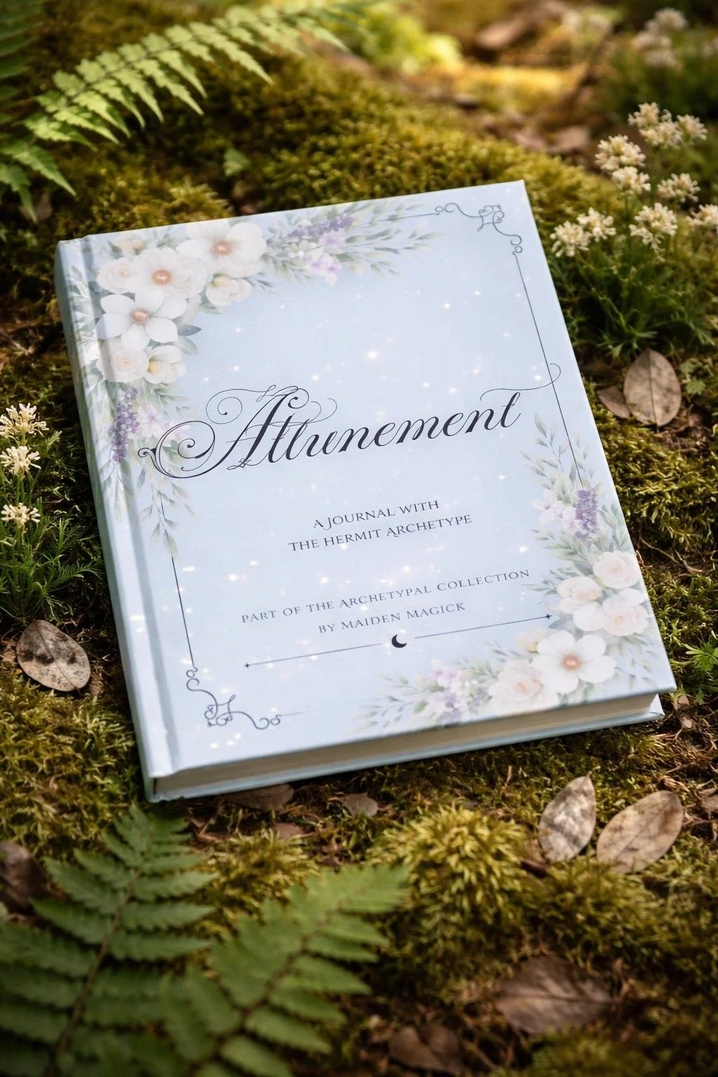 A book titled 'Attunement: A Journal with the Hermit Archetype' lies on the mossy ground surrounded by ferns and small plants in a natural outdoor setting.