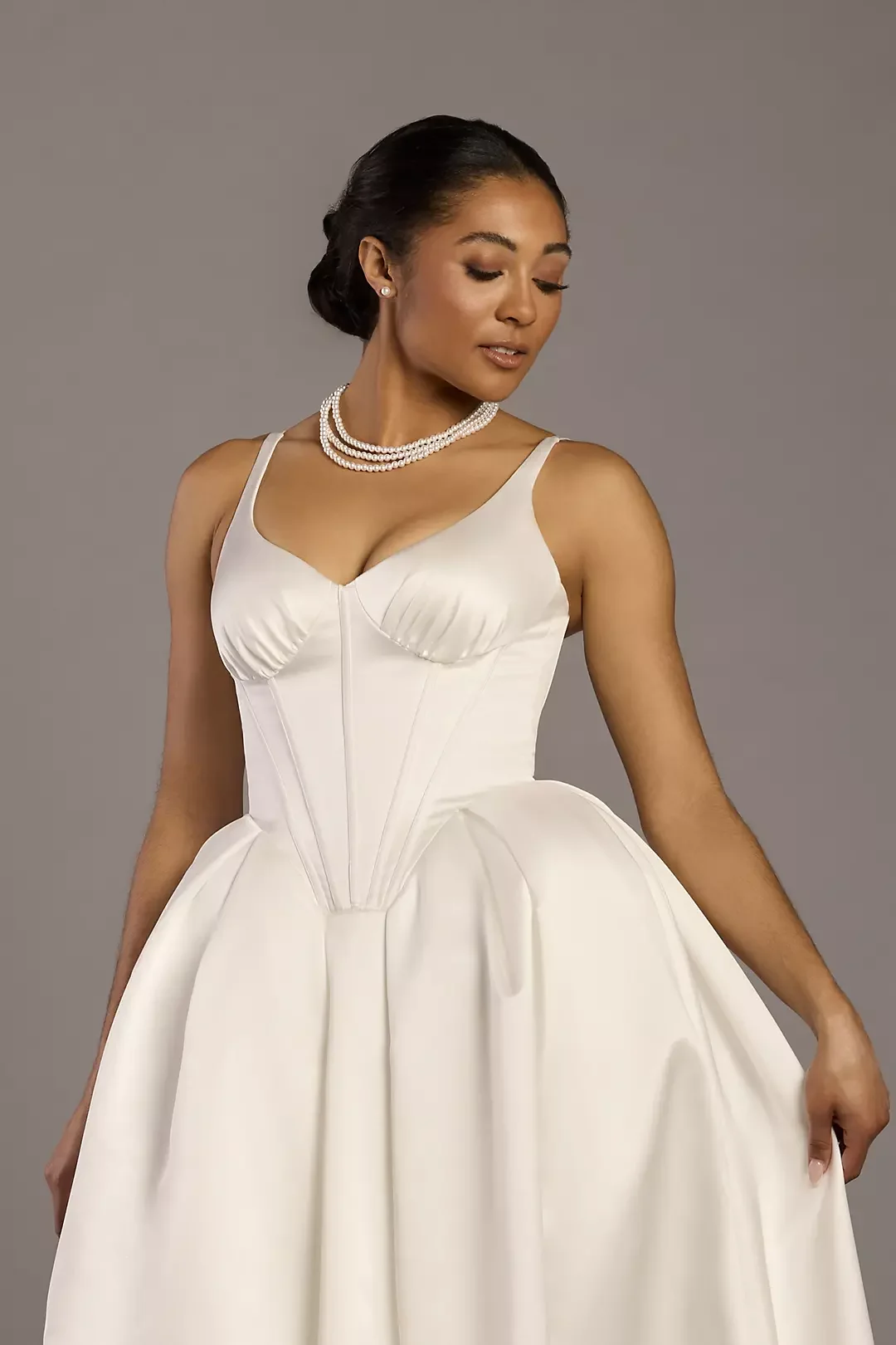 Woman in a white gown with a sweetheart neckline, pearl jewelry, and a sophisticated updo hairstyle, standing against a plain gray background.