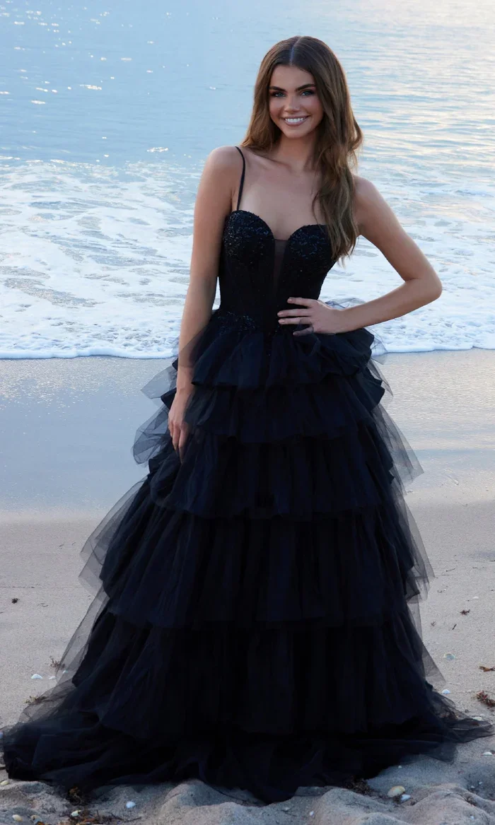 A woman in a black ball gown standing on a beach with ocean waves in the background.