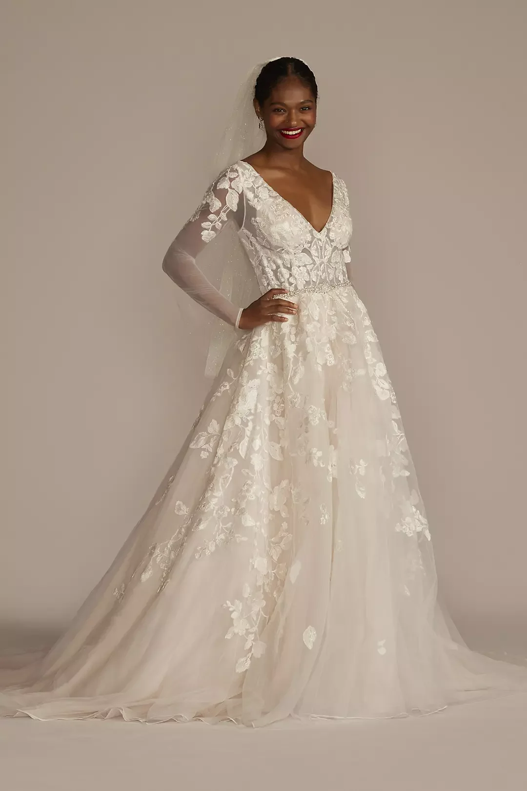 Woman in a white wedding gown with floral lace details, long sheer sleeves, and a veil, standing against a plain neutral background.