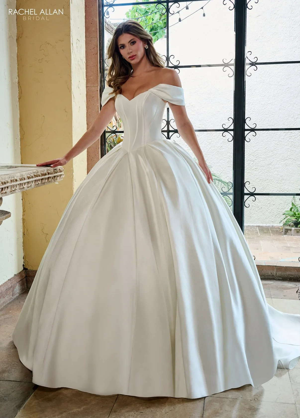 A woman wearing an off-the-shoulder white wedding gown, standing indoors near a window with iron bars, with her left hand resting on a stone ledge.