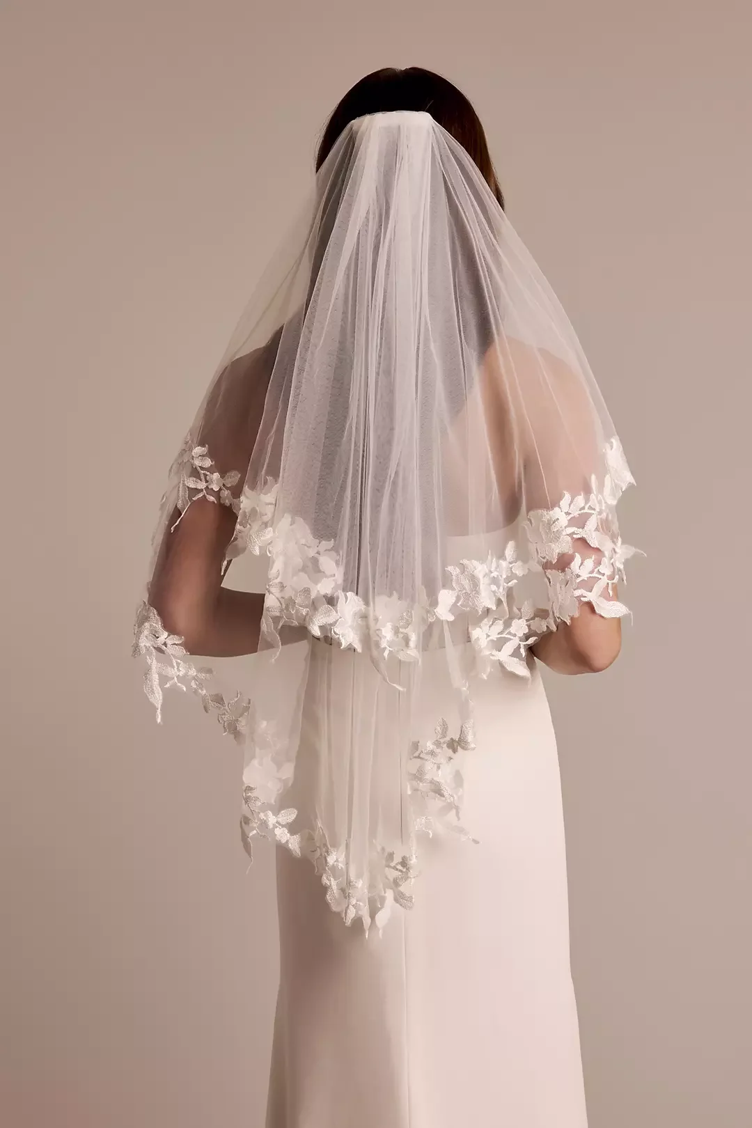 Back view of a bride in a white wedding dress with an off-shoulder lace detail and a long veil with floral embroidery, standing against a neutral background.