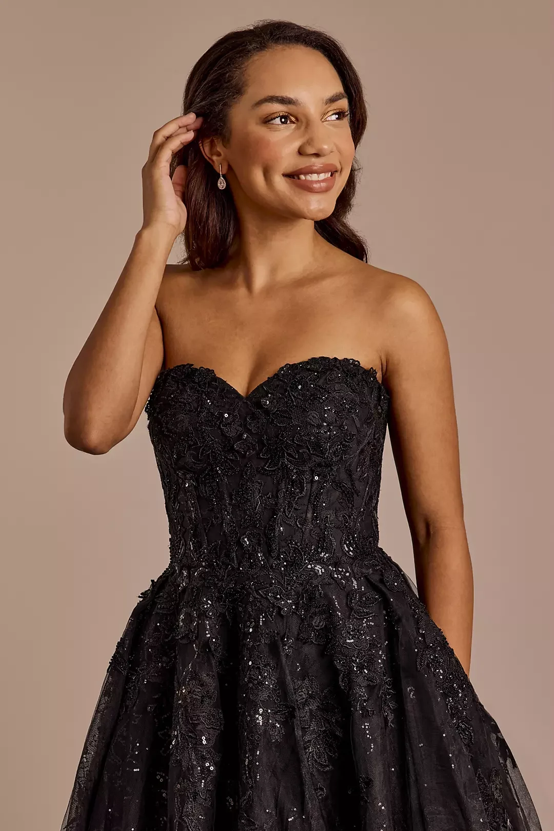 A woman in a strapless black lace evening gown with sequins, smiling, touching her hair, with earrings, against a plain beige background.