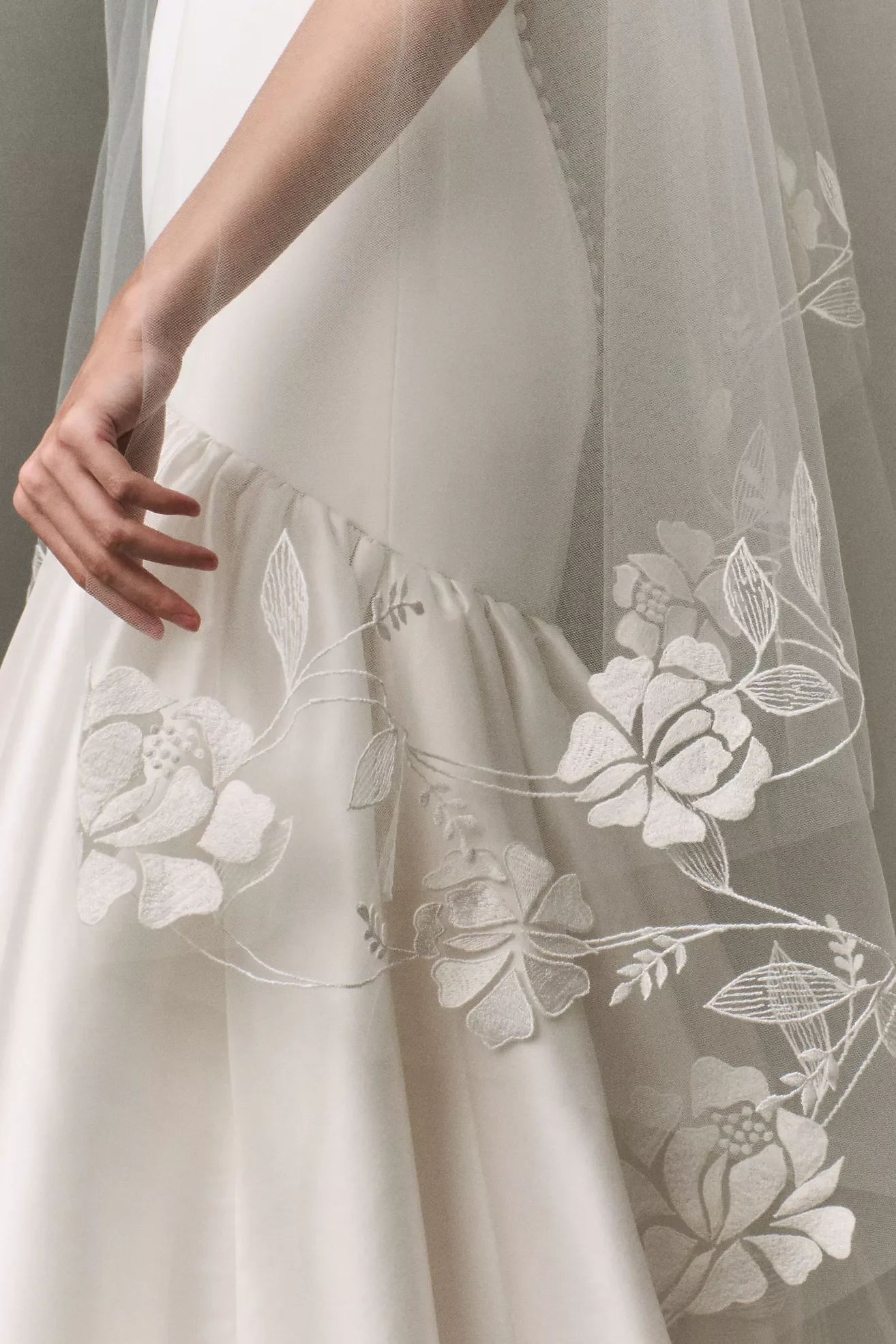 Close-up of a hand touching the hem of a white lace wedding dress with floral embroidery.