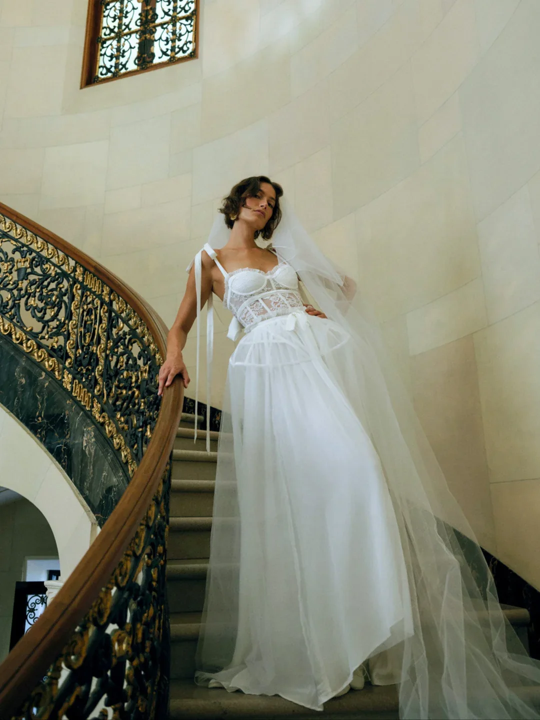 A woman in a white, lace, and tulle wedding dress with a veil stands on a curved staircase with ornate black and gold railing, in a beige wall interior.