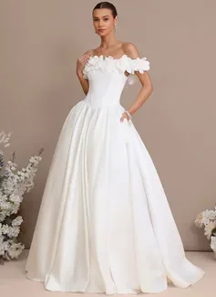 Woman in an off-shoulder white wedding dress with floral appliqué details, standing indoors with white flowers in the background.