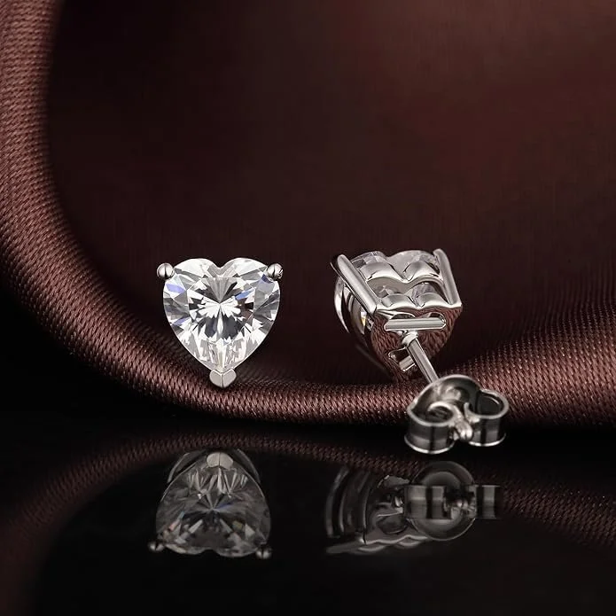 A pair of silver heart-shaped diamond stud earrings resting on a dark brown satin fabric.