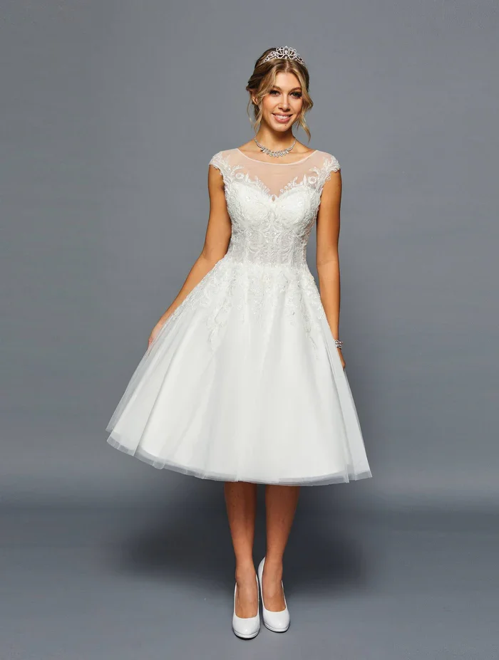 A woman in a white wedding dress with lace details, wearing a tiara and jewelry, standing against a plain gray background.
