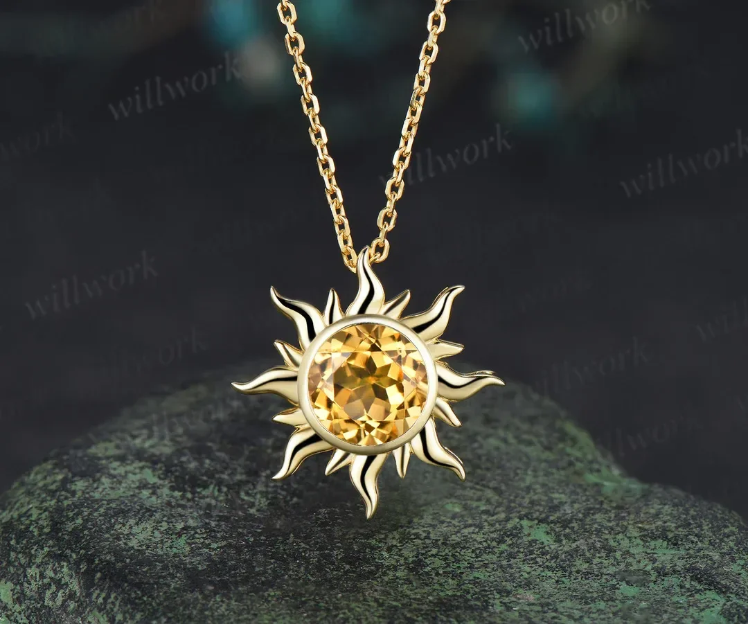 Gold sun-shaped necklace with a large amber-colored gemstone in the center, hanging on a chain against a dark background.