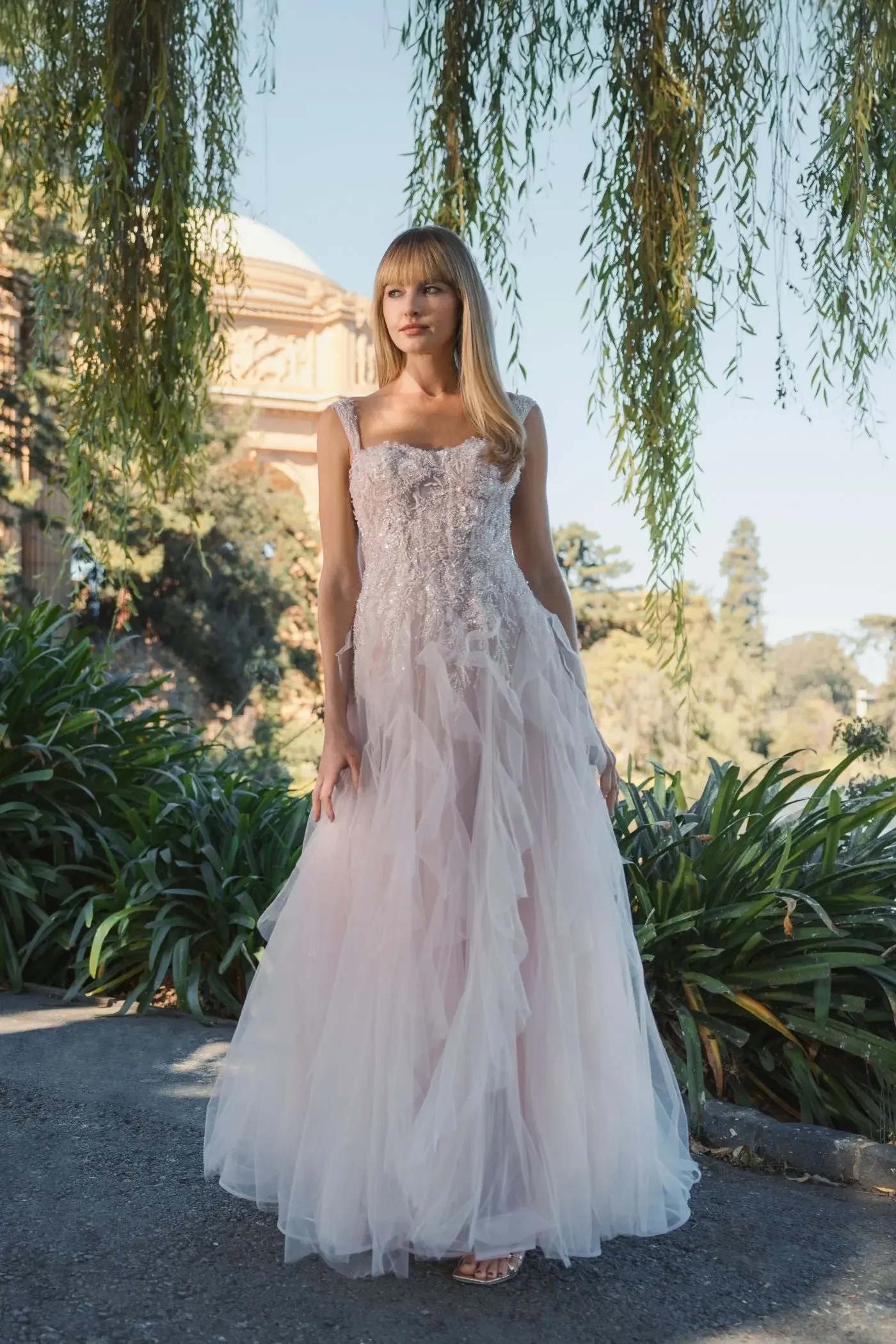 A woman in a white, lace, ball gown style wedding dress standing outdoors under a tree with hanging branches and in front of a building with a dome, surrounded by greenery.