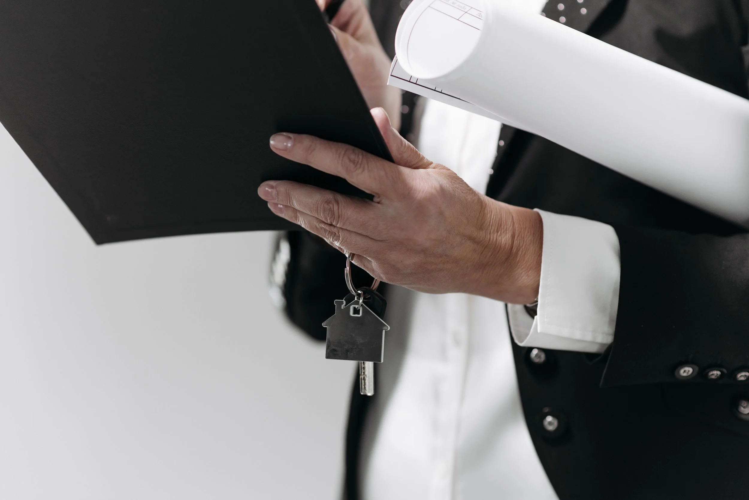 Close-up of a real estate agent holding a black folder, blueprints, and a keychain shaped like a house attached to a key hanging from their hand.