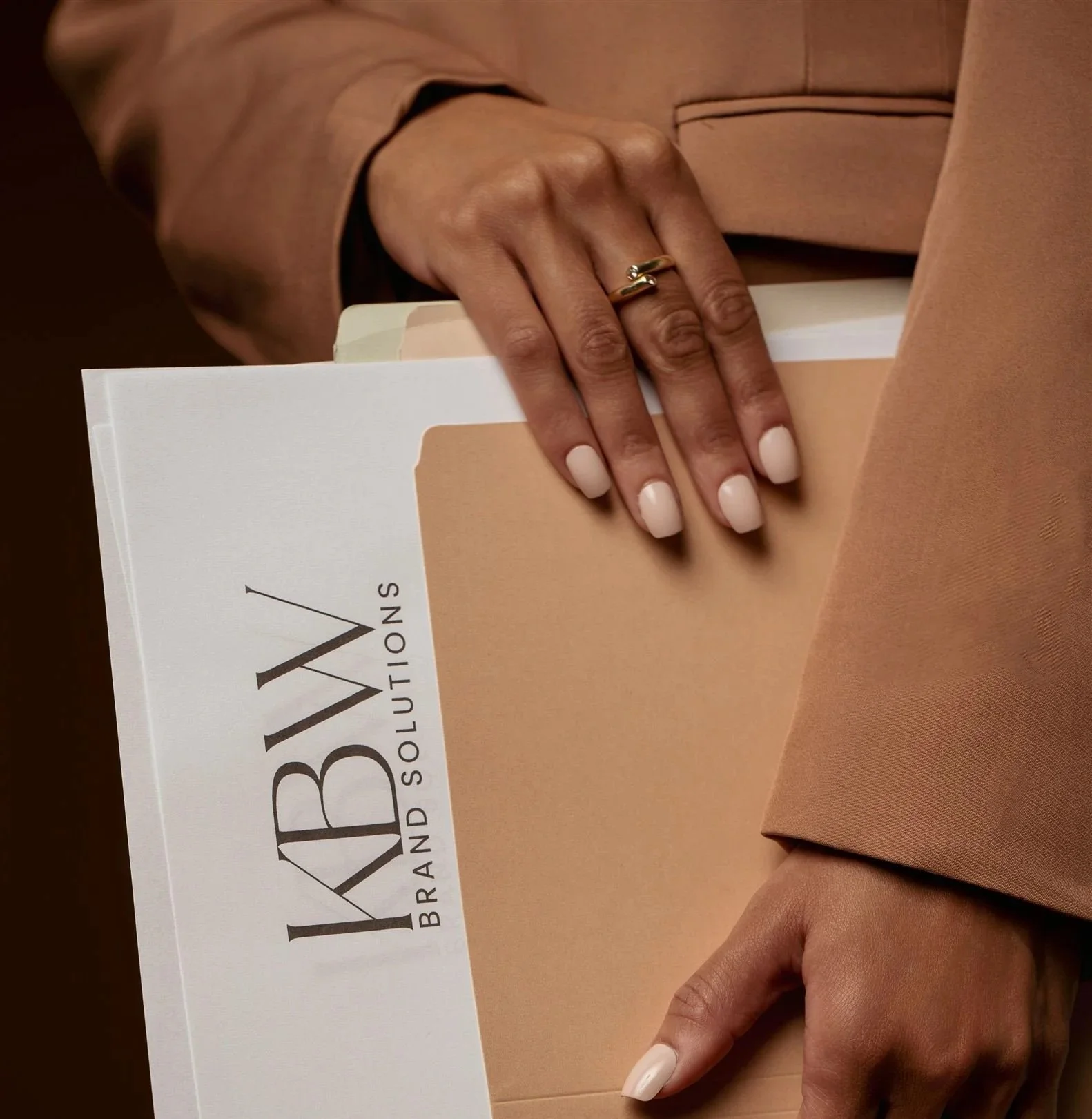 A person's hand with white painted nails holding a beige folder with text, wearing a gold ring and a beige blazer.