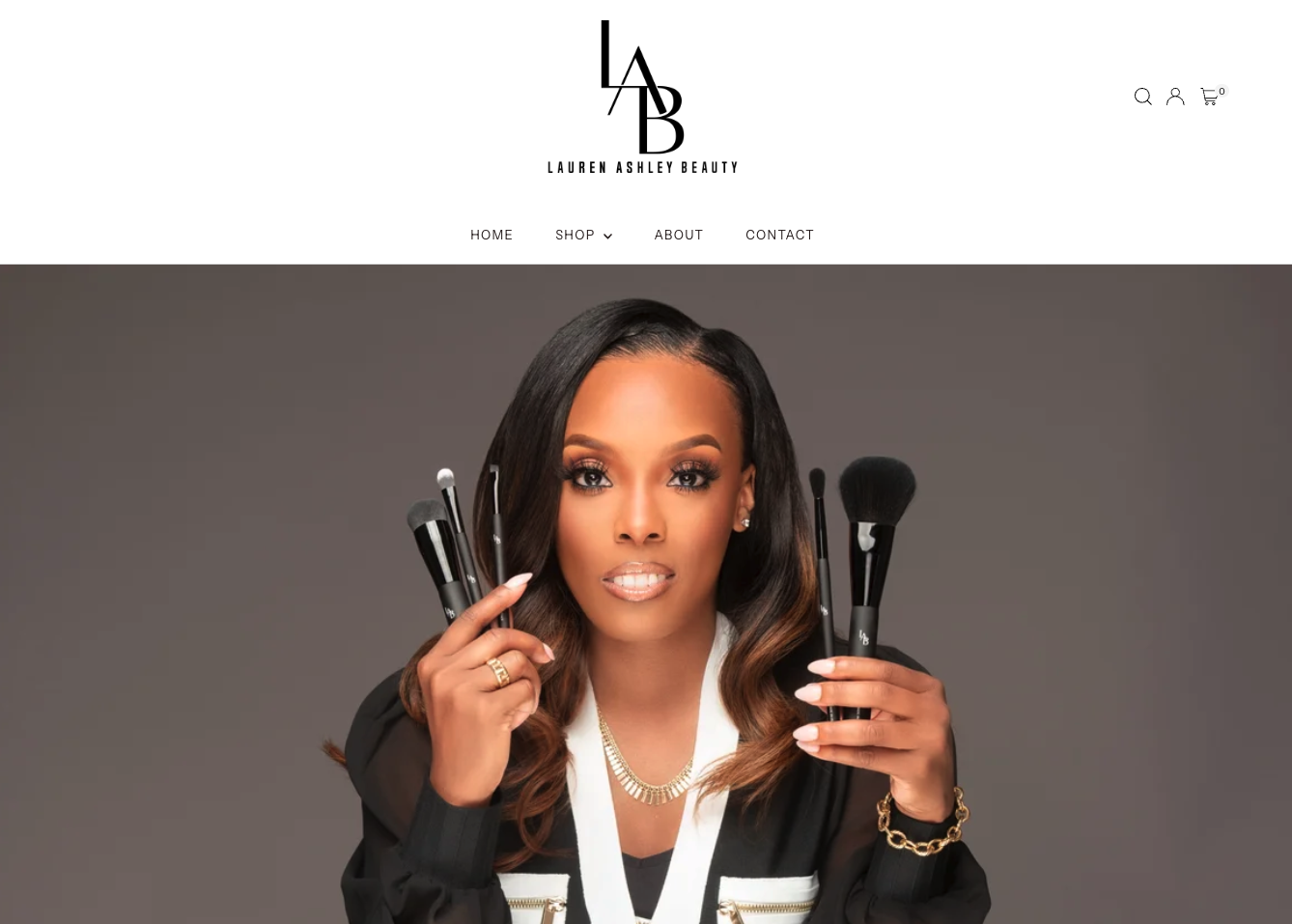Professional makeup artist and businesswoman, Lauren Ashley, with makeup brushes posing for promotional photo on her beauty brand's website, holding makeup brushes with a neutral gray background, wearing a black and white outfit and gold jewelry.