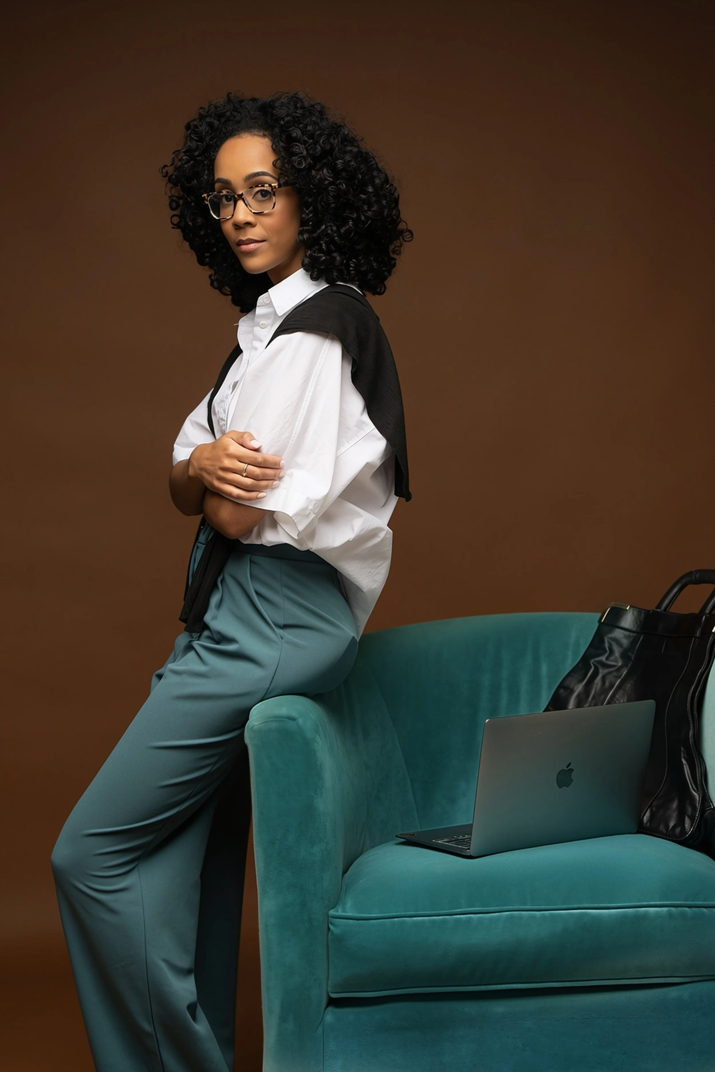 KBW Brand Solutions founder, Katie Woods, with curly black hair, glasses, and a white shirt sits on an emerald green velvet armchair, with a laptop and a black handbag nearby. The background is brown.