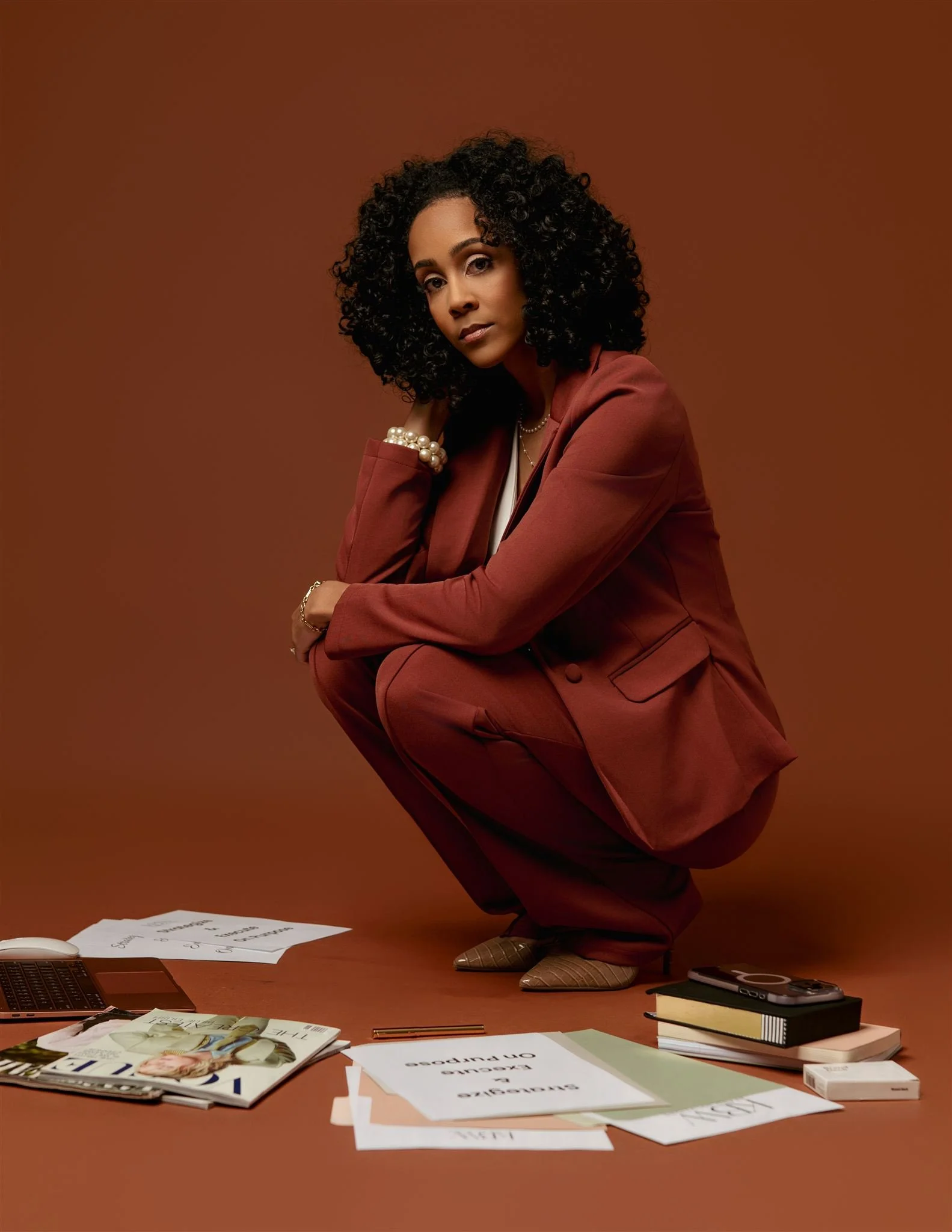 KBW Brand Solutions founder, Katie Woods, with curly black hair and light brown skin, dressed in a brown suit, squatting on a brown floor with papers, magazines, books, a calculator, and a phone scattered around with a brown background.