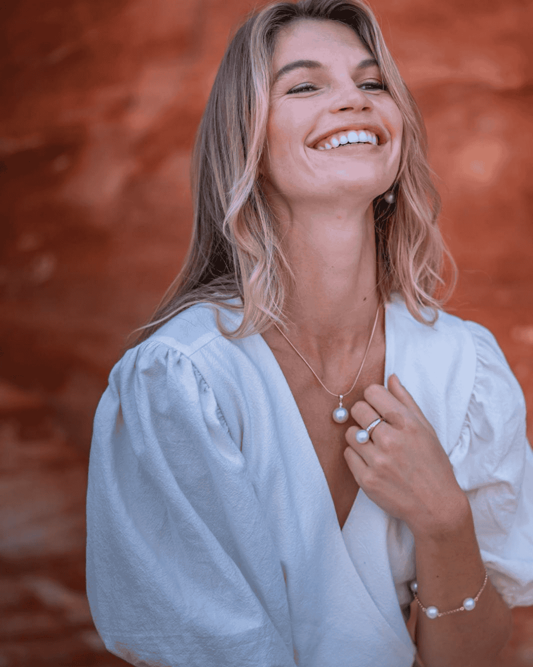 A smiling woman with wavy blonde hair wearing pearl jewelry and a white blouse, standing outdoors with a blurred reddish-brown background.