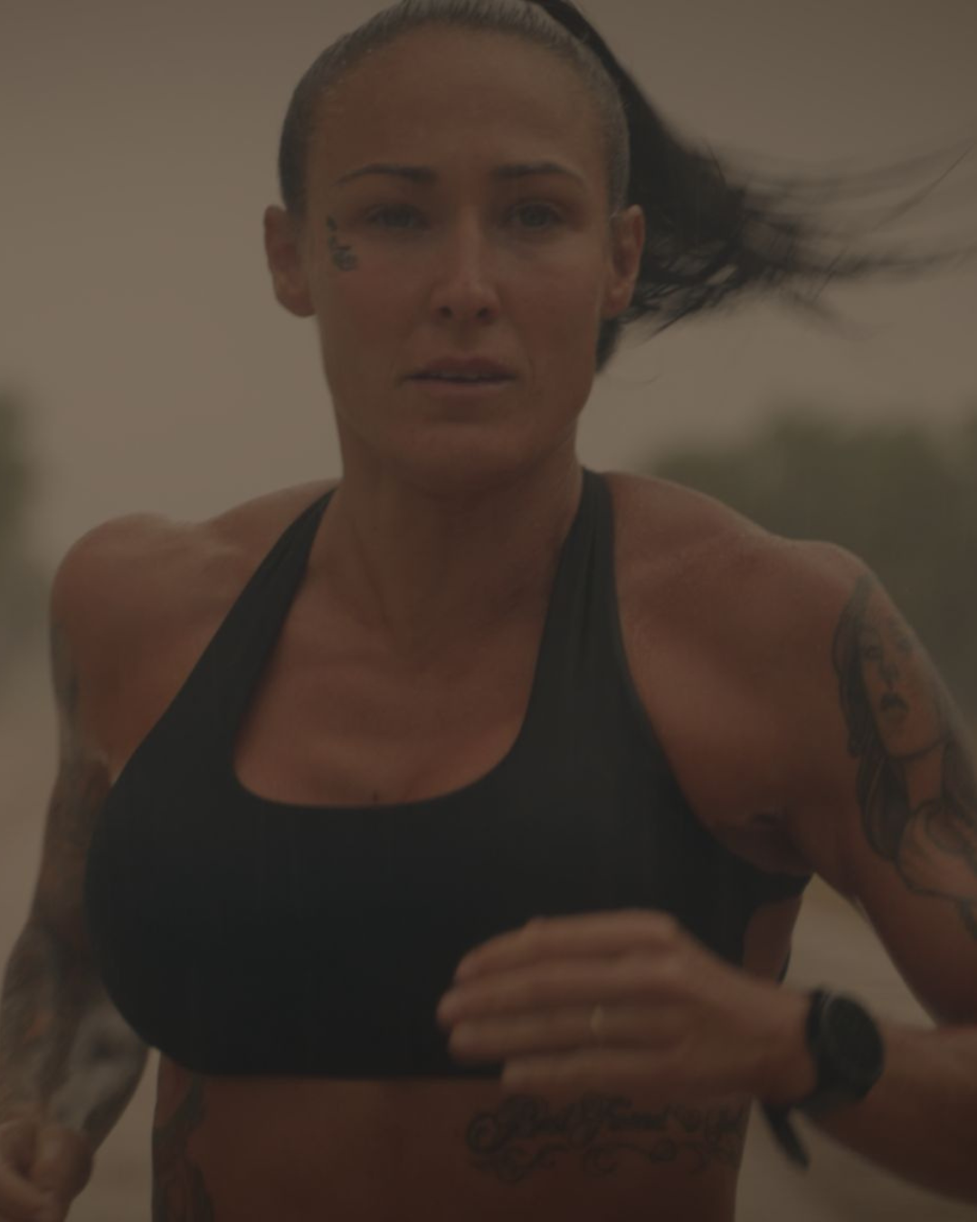 A woman with dark hair running outdoors. She has multiple tattoos on her arms and chest, and is wearing a black sports bra and a wristwatch.