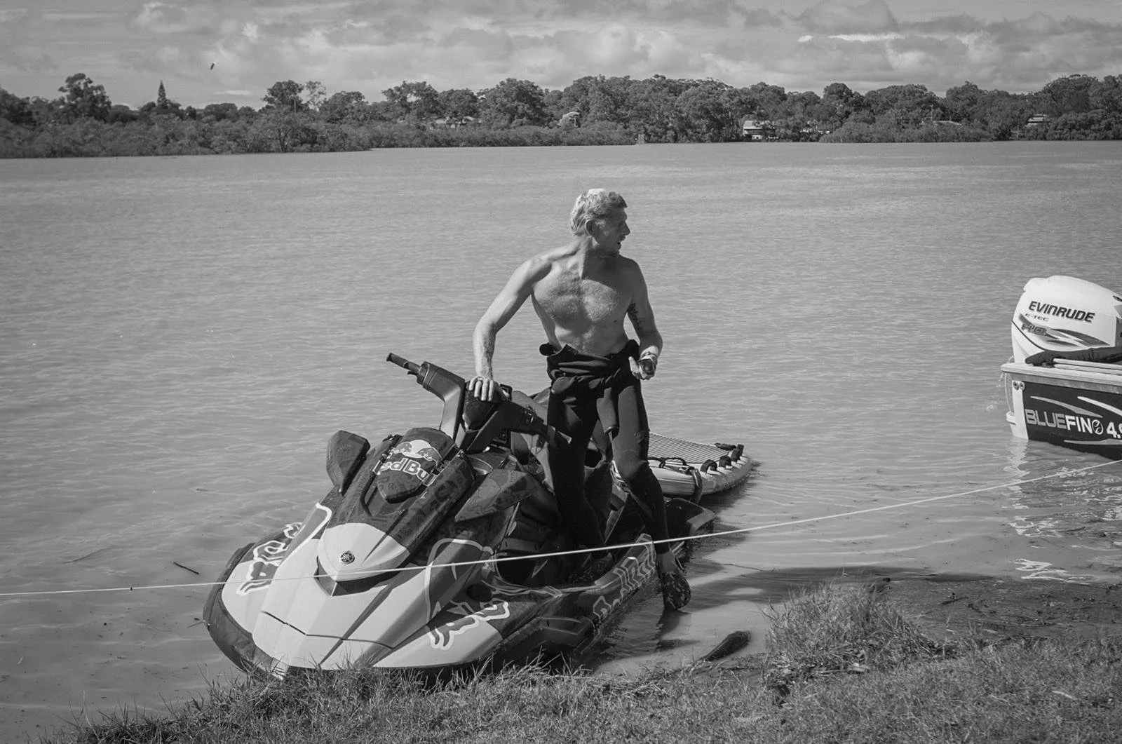 A shirtless man with gray hair standing on a jet ski at the edge of a lake, holding a small object in his right hand, with a boat tied to the jet ski. Trees and houses are visible in the background.