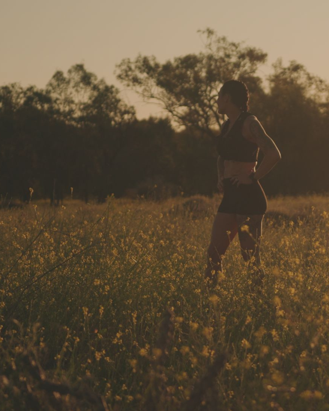 Silhouette of a woman jogging through a field of yellow flowers during sunset with trees in the background.