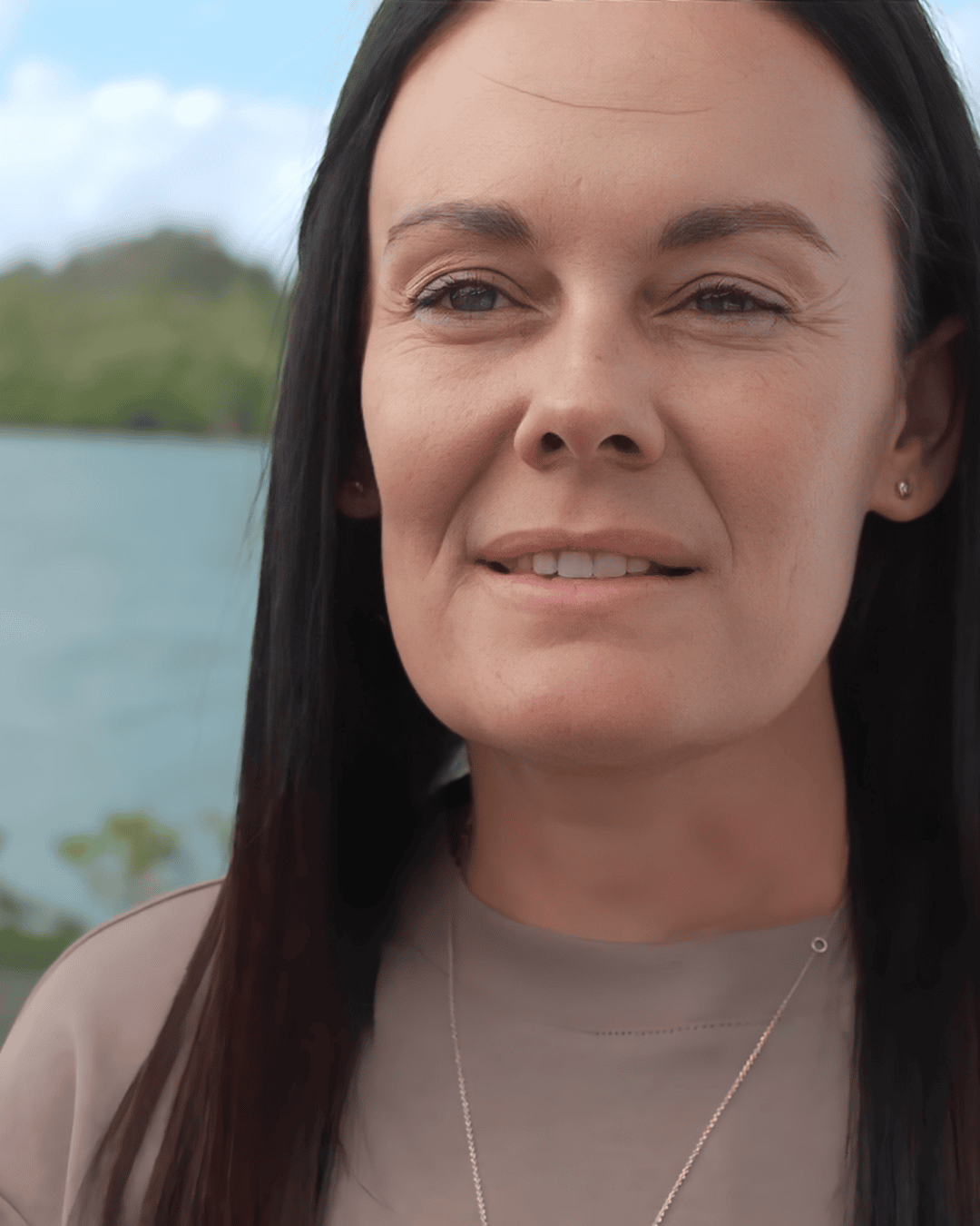 Close-up of a woman with long dark hair, light skin, and a subtle smile, standing outdoors near water with trees in the background.