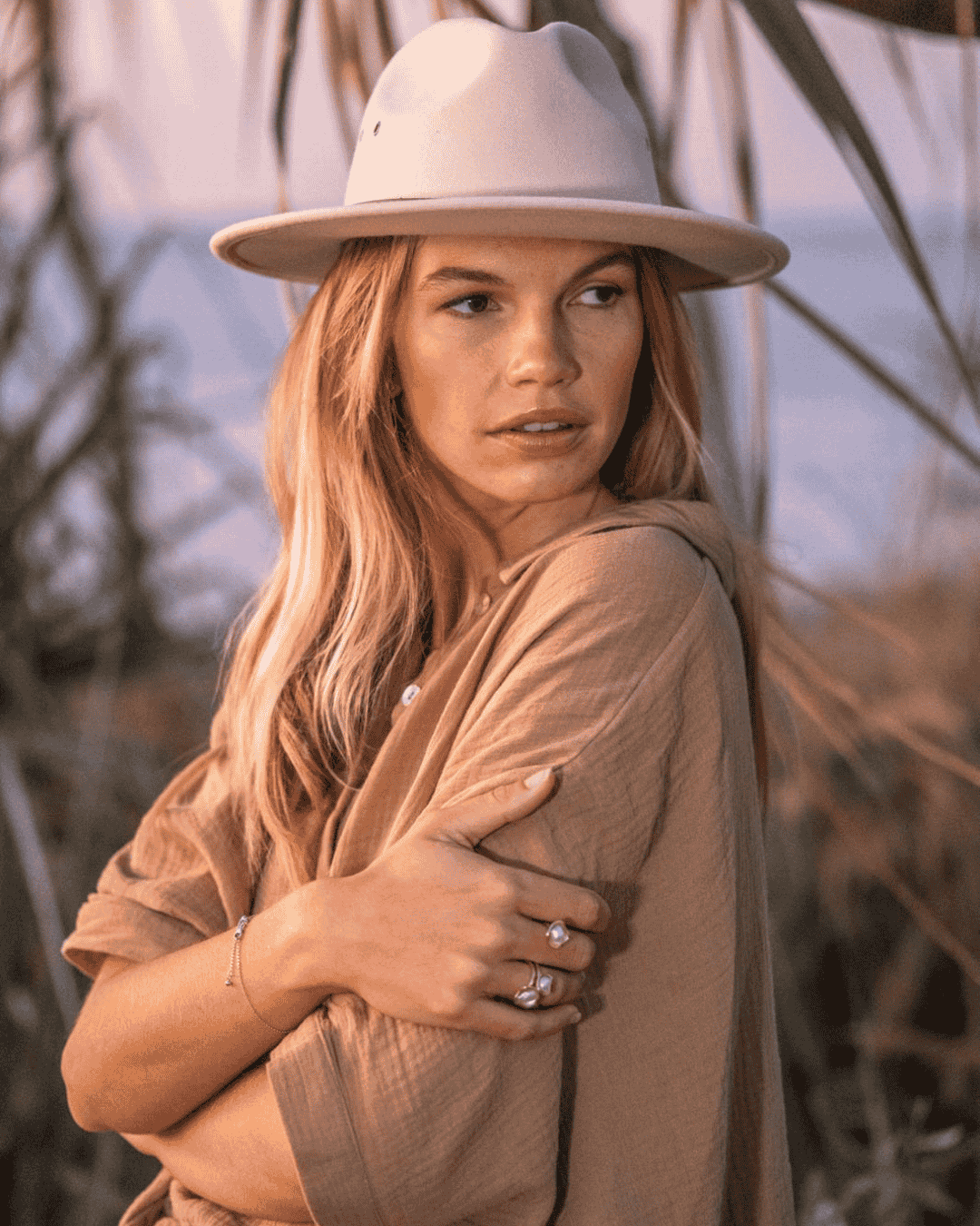 A woman with wavy, blonde hair wearing a beige wide-brimmed hat and a beige shirt, standing outdoors near tall grasses, with a calm expression and arms crossed.