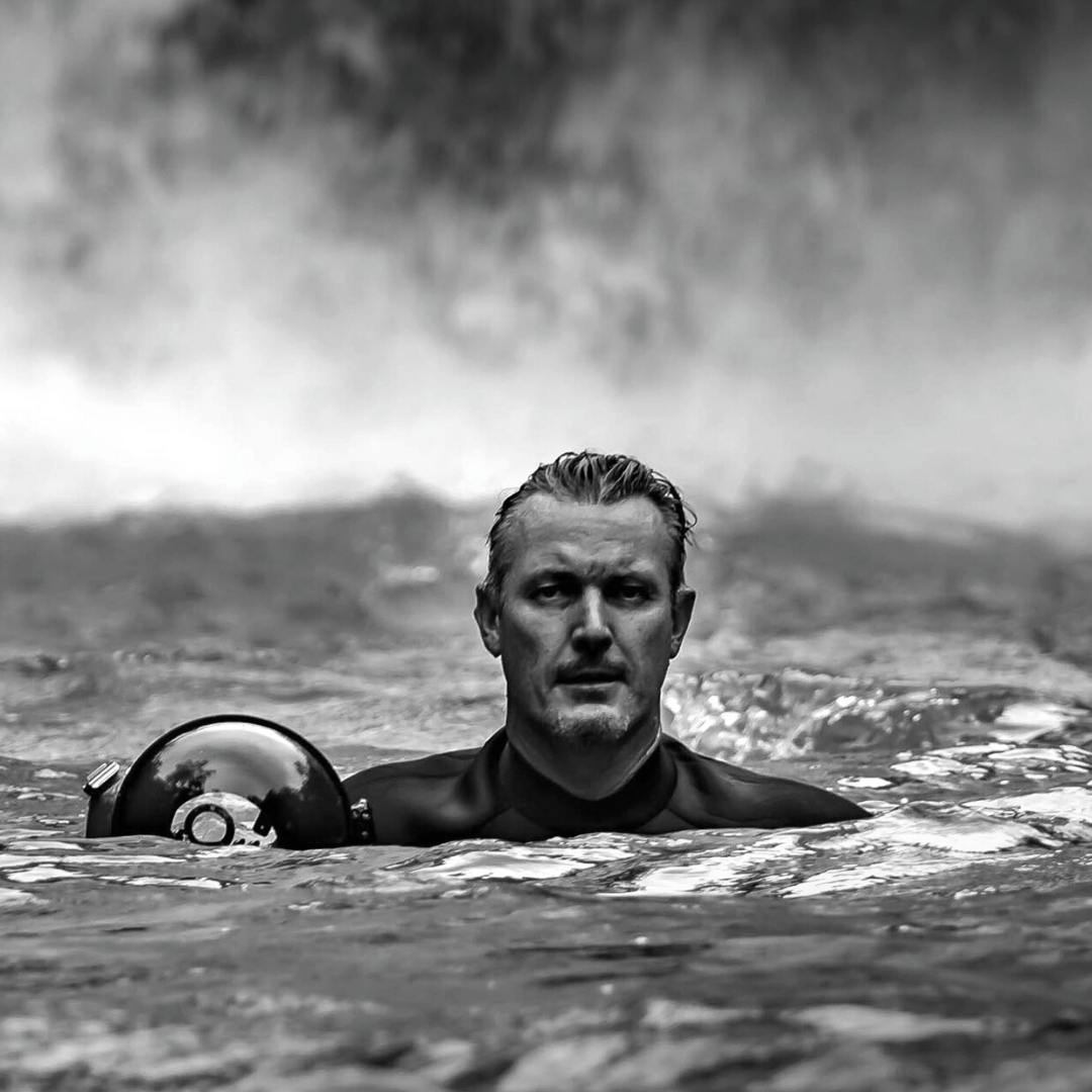 Black and white photo of a award winning director Stuart McKay in water with a serious expression, holding a helmet, with a waterfall in the background.