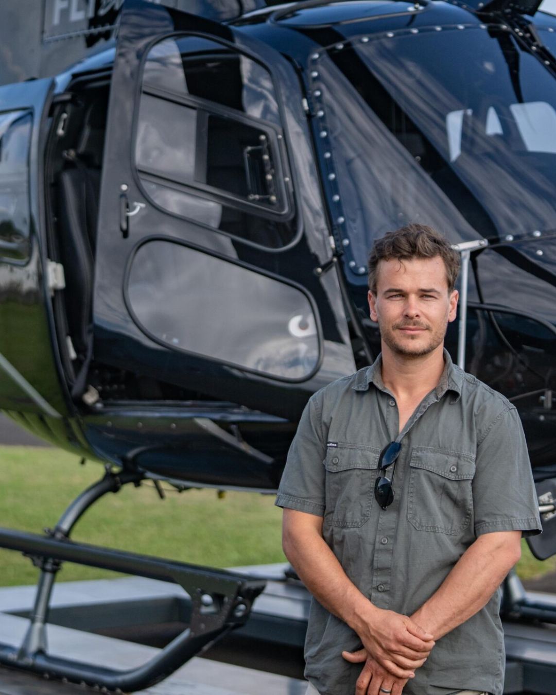 A man with sunglasses hanging from his shirt stands in front of a black helicopter.