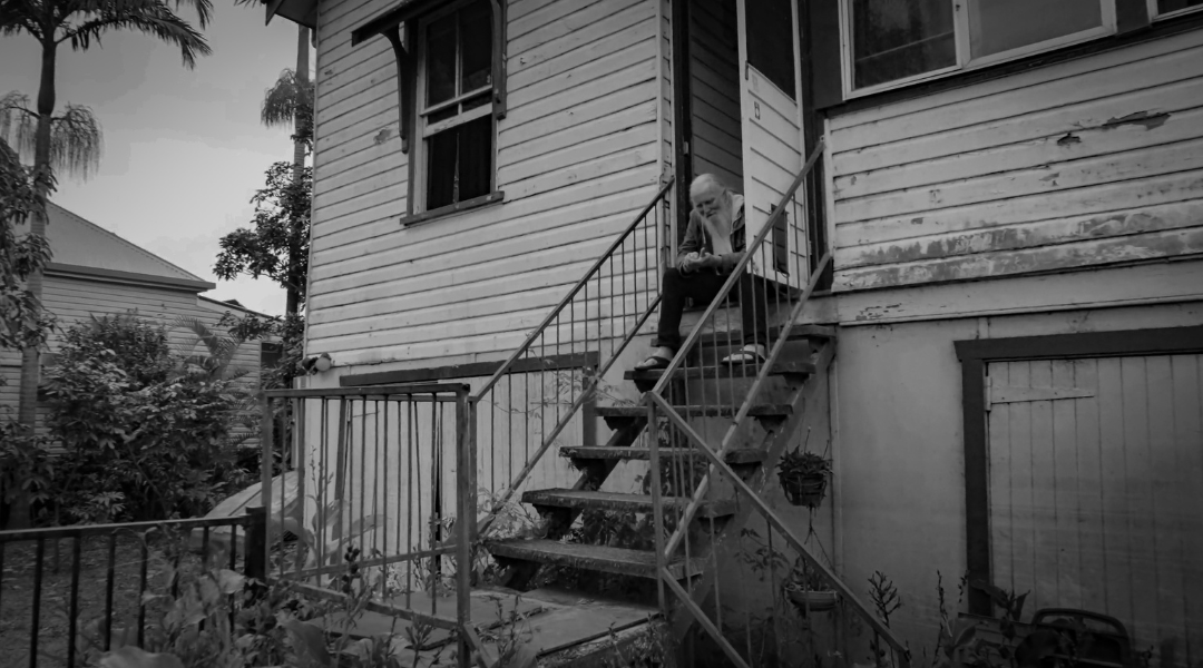 A person sitting on the stairs outside a house, looking down and writing or reading, with trees and another house visible in the background.