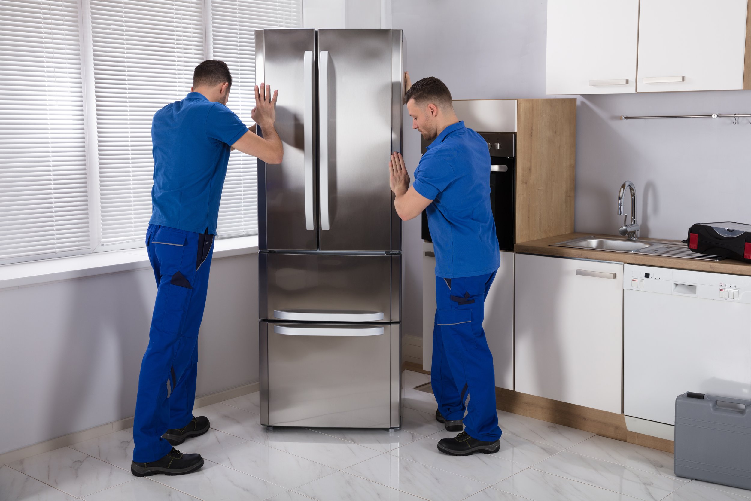 Two technicians in blue uniforms pushing a stainless steel refrigerator in a modern kitchen