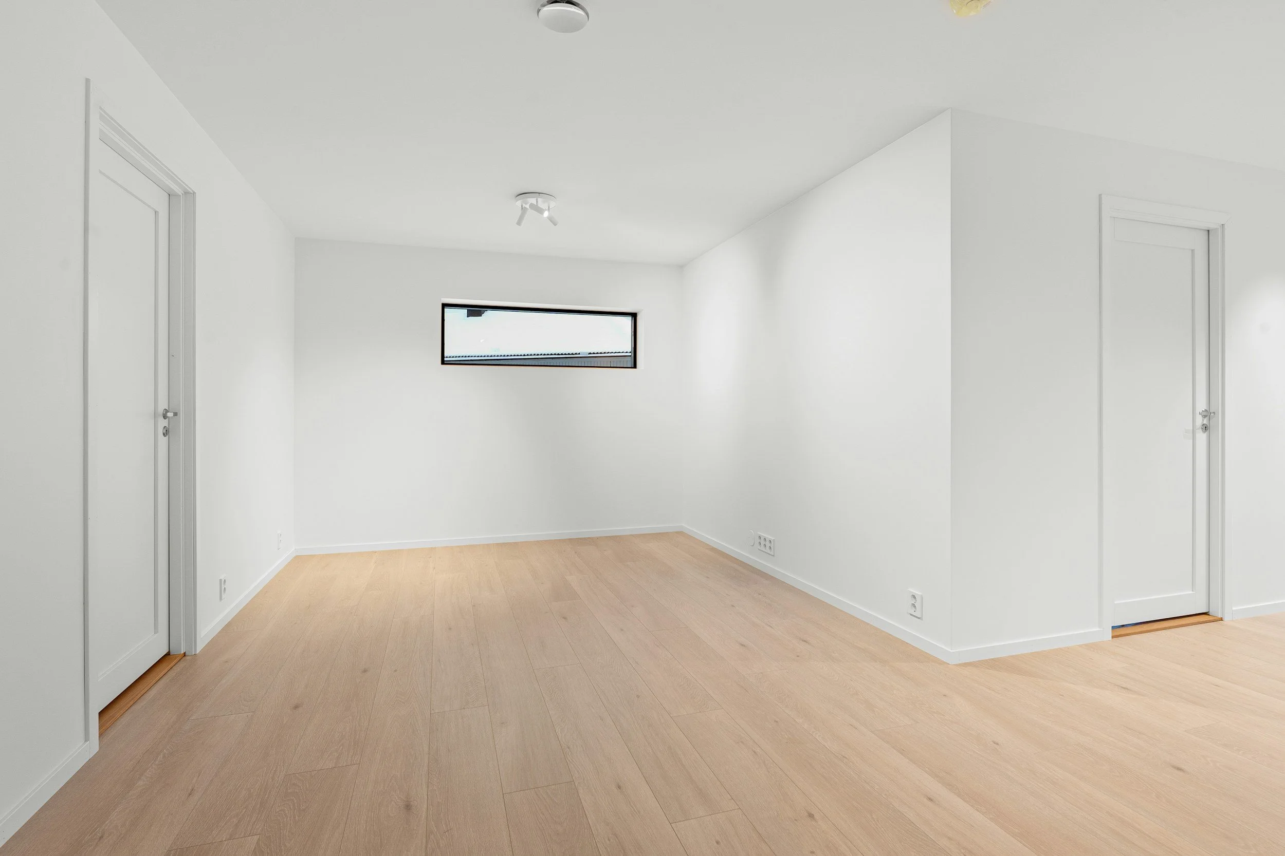 Empty room with white walls, light wood flooring, a small horizontal window, and two closed doors.