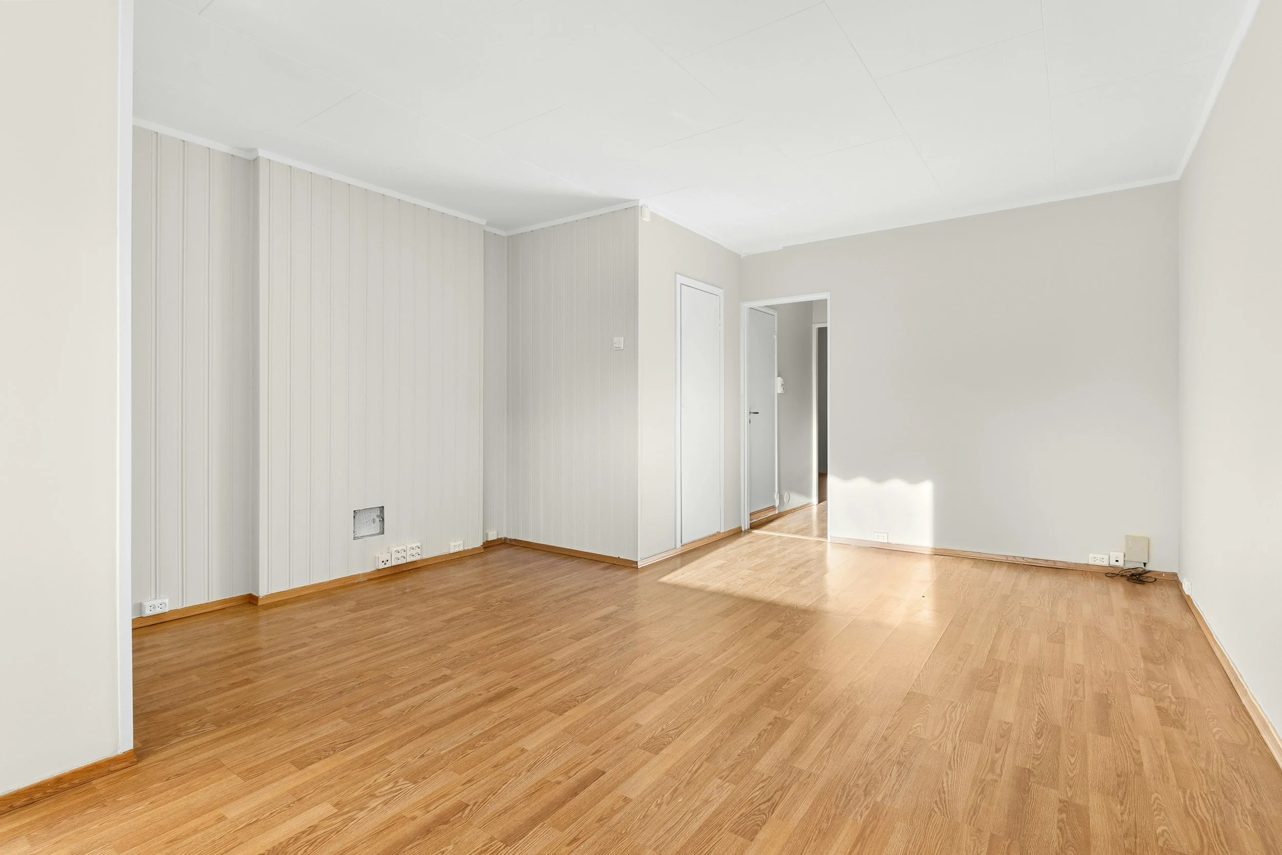 Empty living room with white walls, hardwood floors, and a doorway leading to another room.