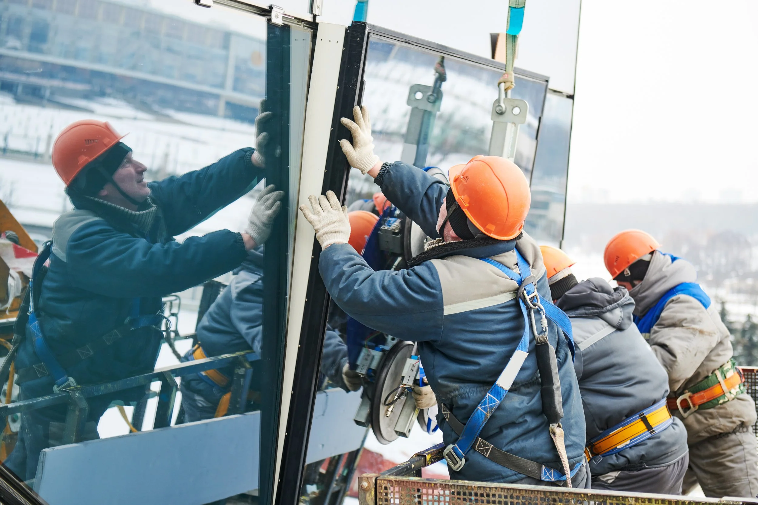 Window installation workers wearing orange safety helmets and harnesses installing a large glass window outdoors.