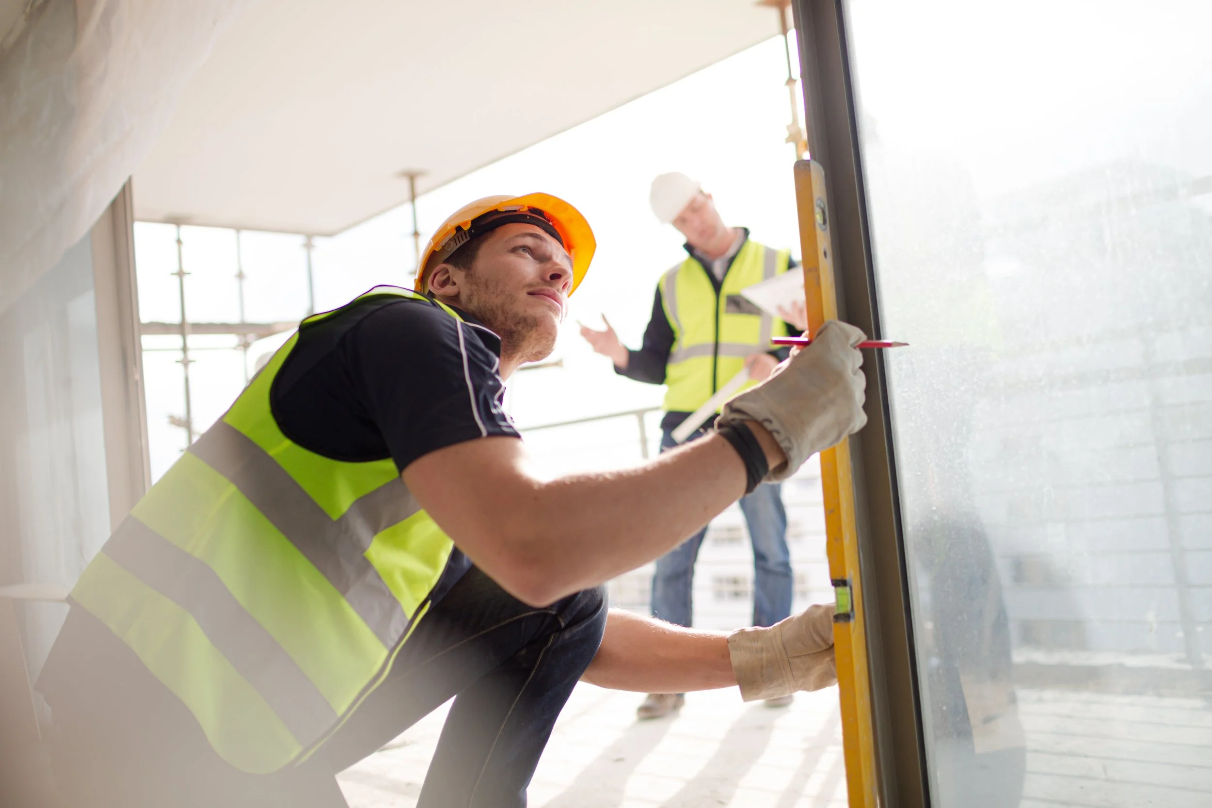 A construction worker in a yellow safety vest and orange helmet kneels down to measure a glass window with a level tool, with another worker in the background reviewing plans on a construction site.