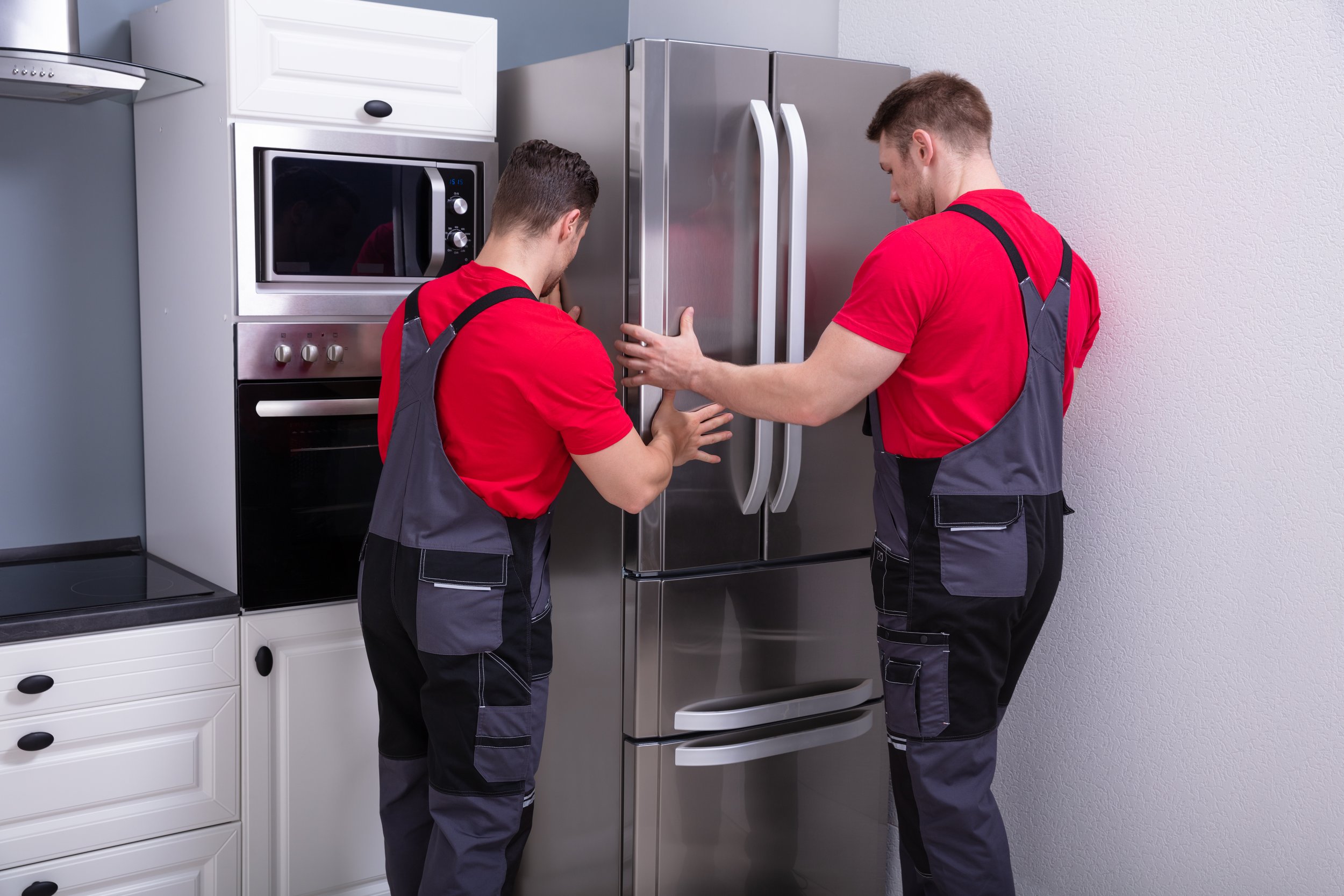 Two workers in red shirts and gray work overalls installing or repairing a stainless steel refrigerator in a kitchen.