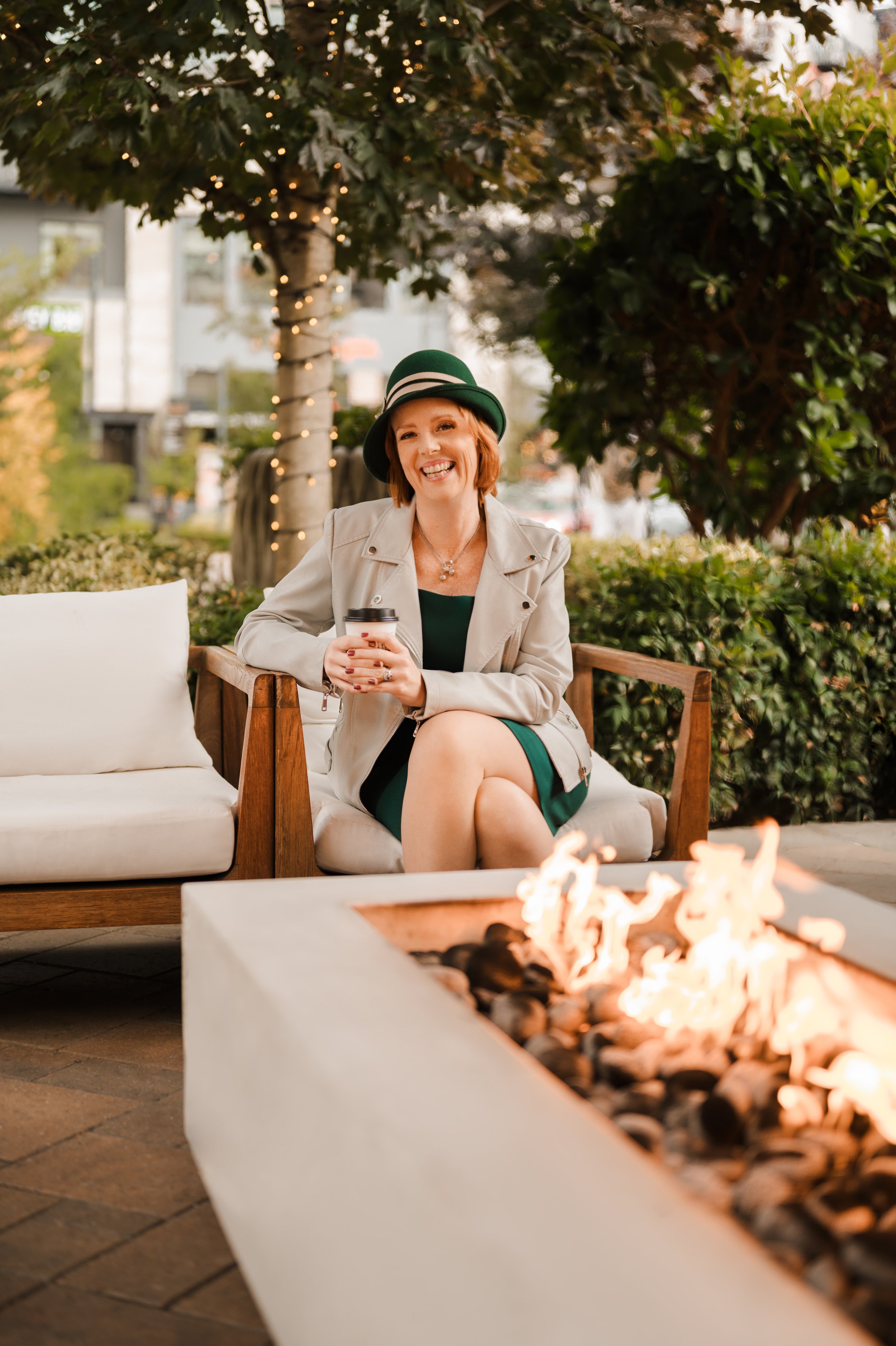 A woman with red hair, wearing a green hat and coat, sitting on an outdoor beige sofa near a fire pit, smiling and holding a cup in her hands.